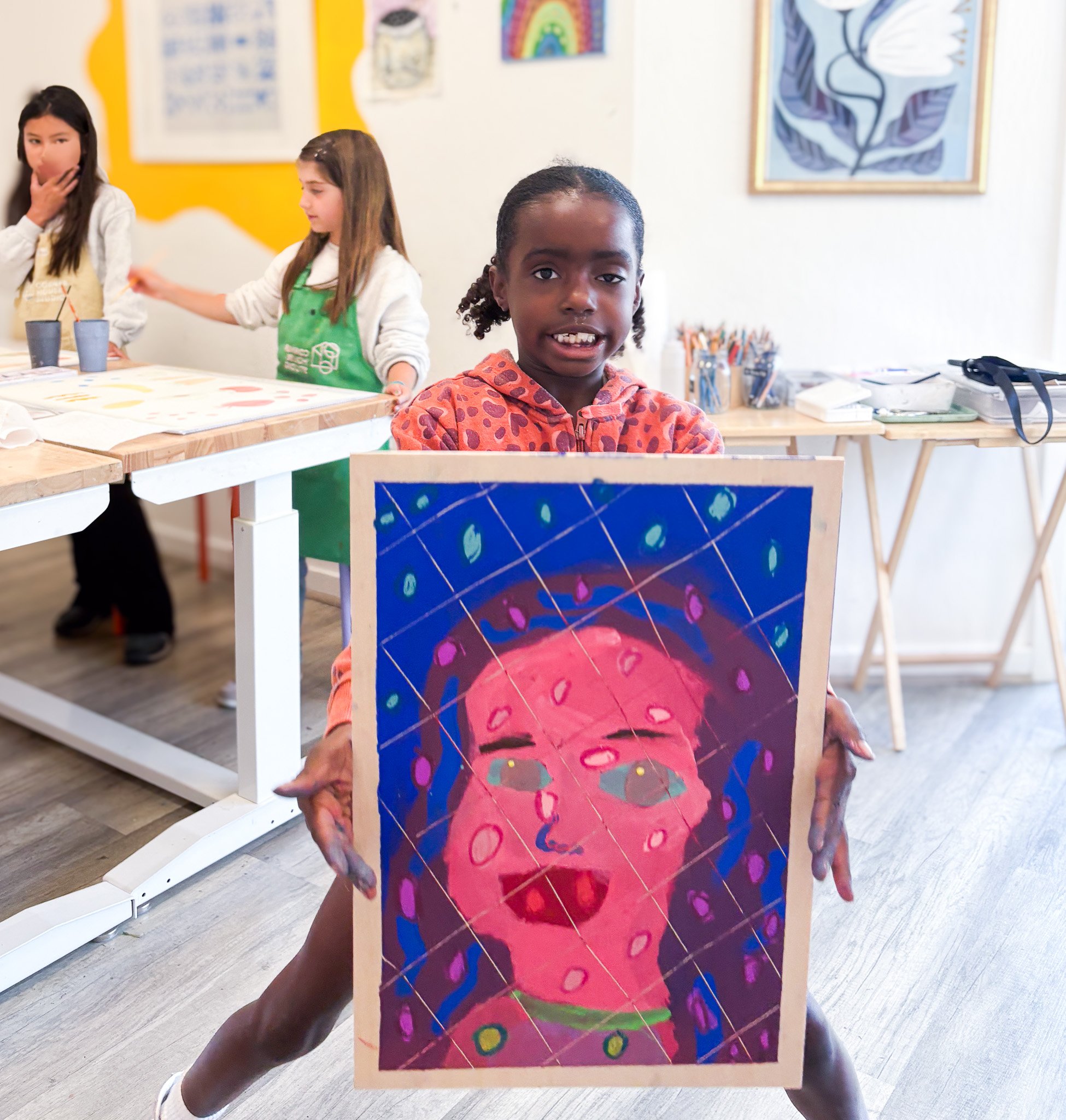 A young girl holding a colorful abstract portrait painting in an art classroom. She is wearing a pink and orange hoodie and smiling at the camera. There are two other children in the background working on art projects at tables.
