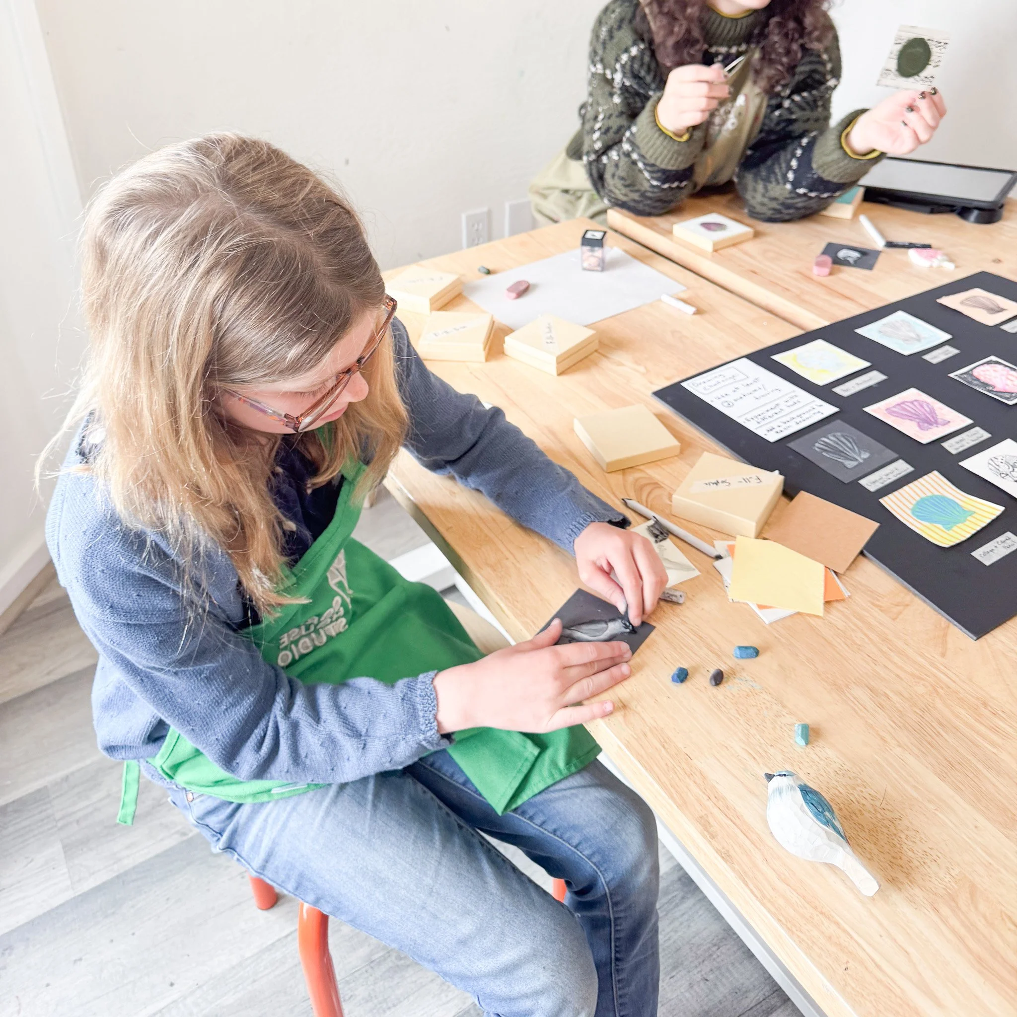 A woman in a green apron working on an art project with small pieces and paper on a wooden table, with another person in the background holding a coin and sitting at the same table.