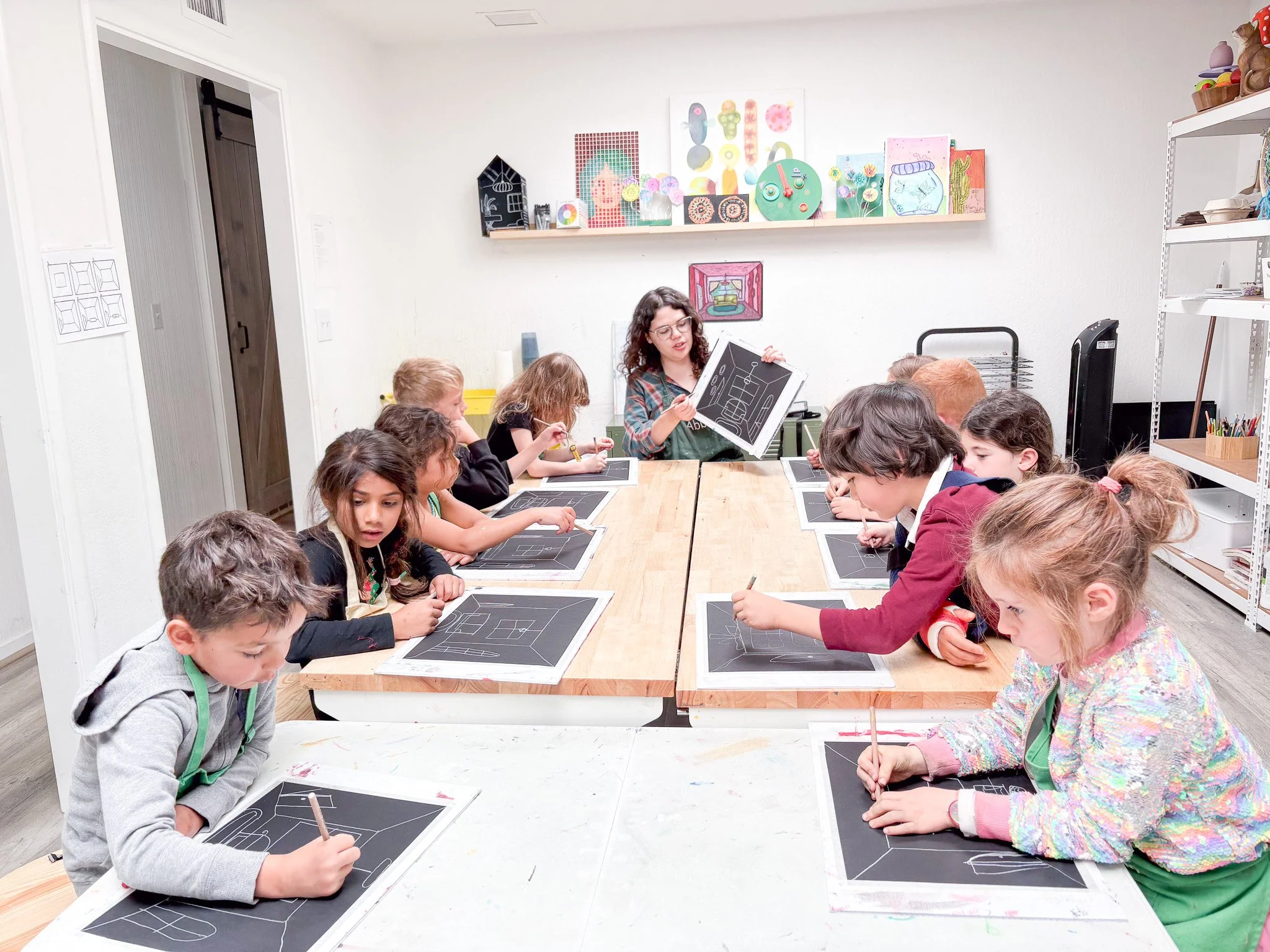 A woman instructs a group of children sitting around a long table, each child working on a chalkboard with chalk, in an art classroom with colorful artwork on the wall.