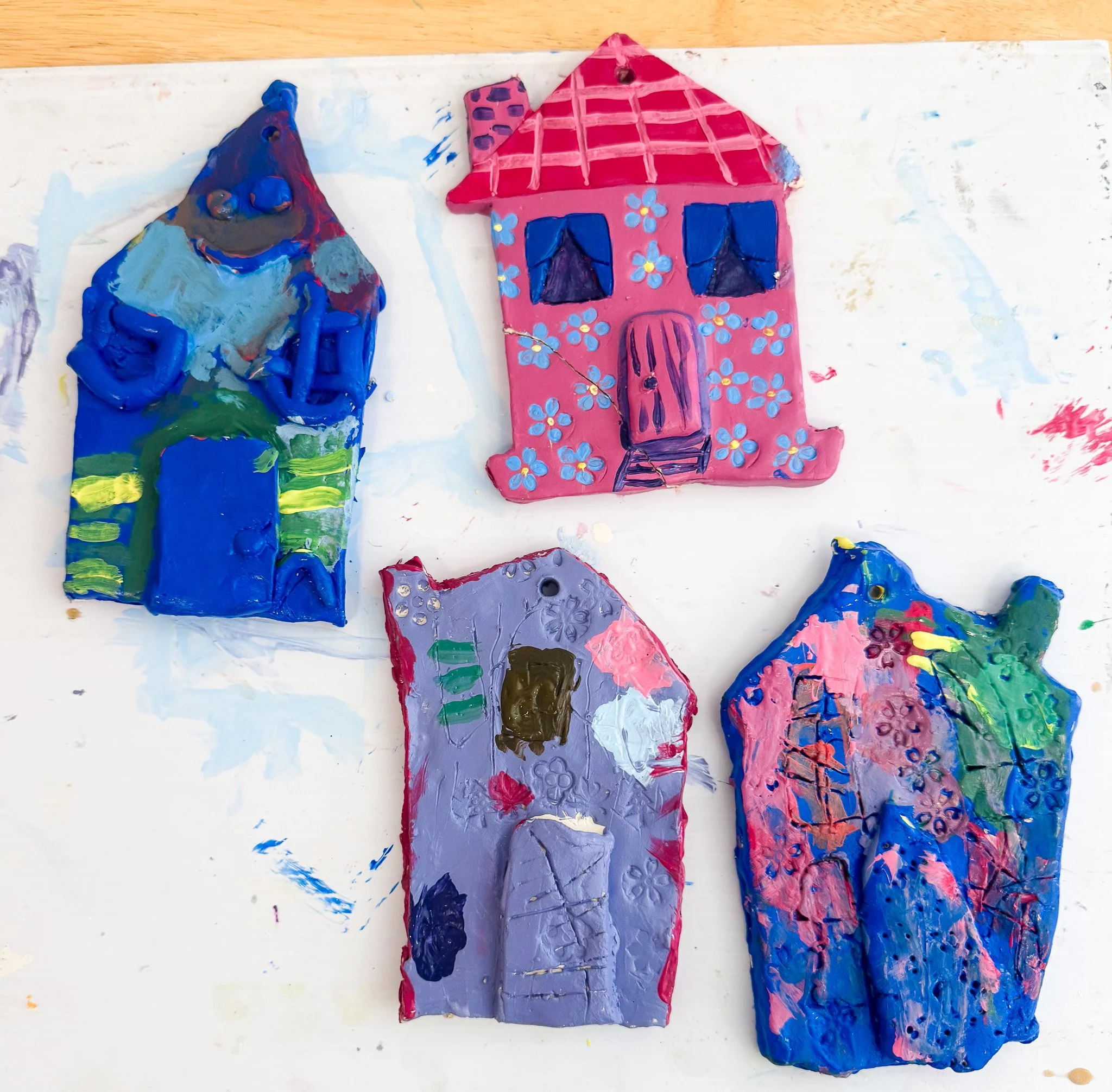 Four colorful painted rocks with childlike house and tree designs on a white surface.