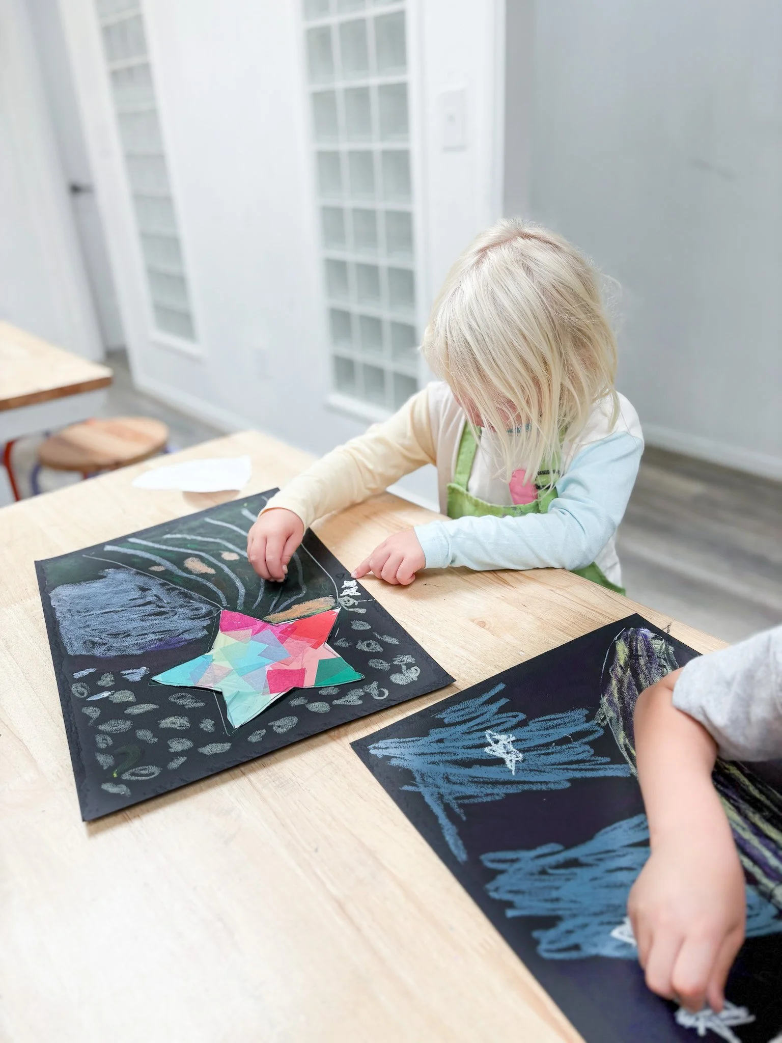 A young girl with blonde hair working on an art project at a wooden table, creating a star-shaped collage on black paper, surrounded by chalk drawings.
