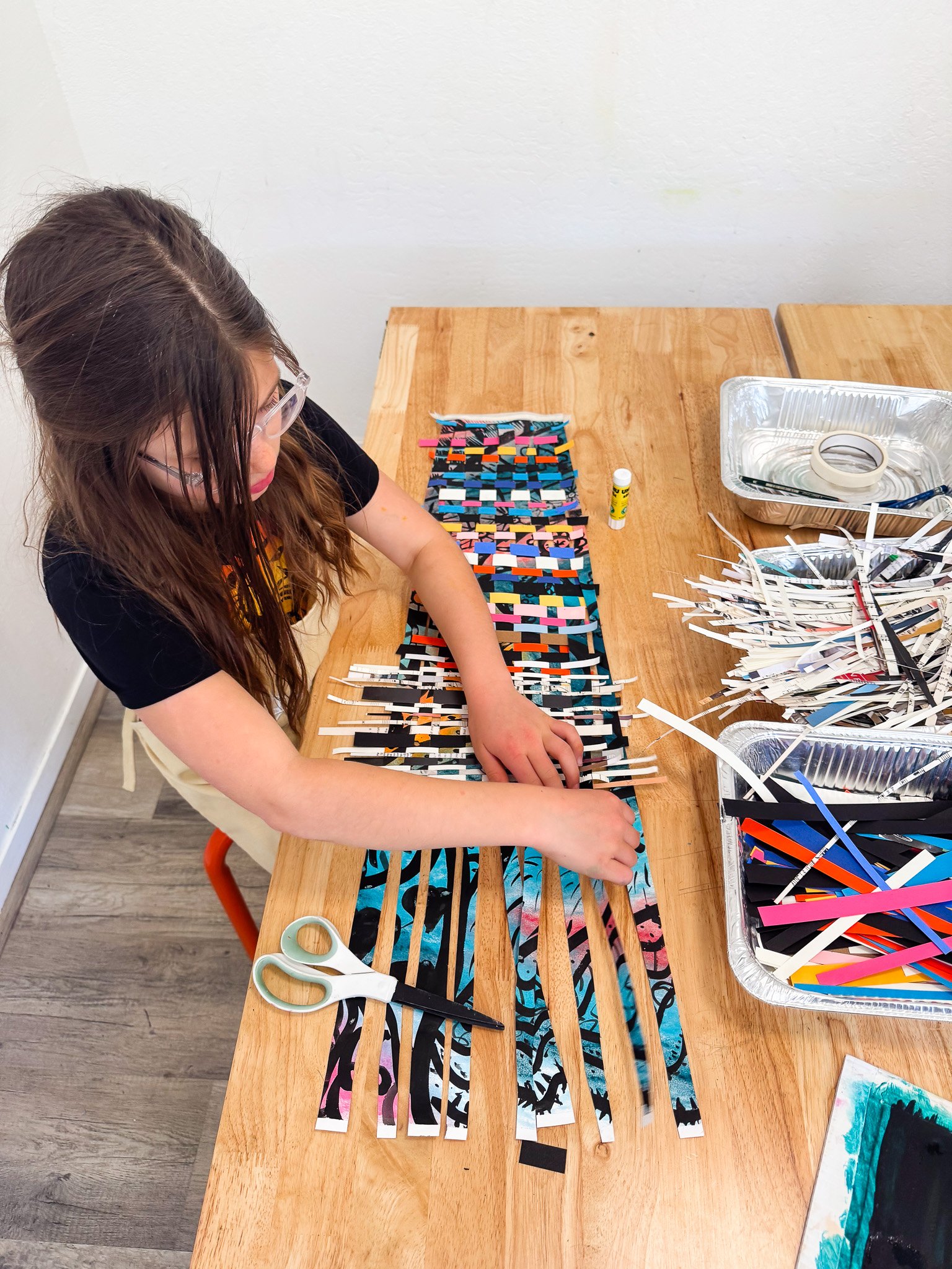 A girl with long brown hair and glasses is creating a paper weaving craft on a wooden table using strips of colorful paper, with scissors, glue, and additional strips of paper nearby.