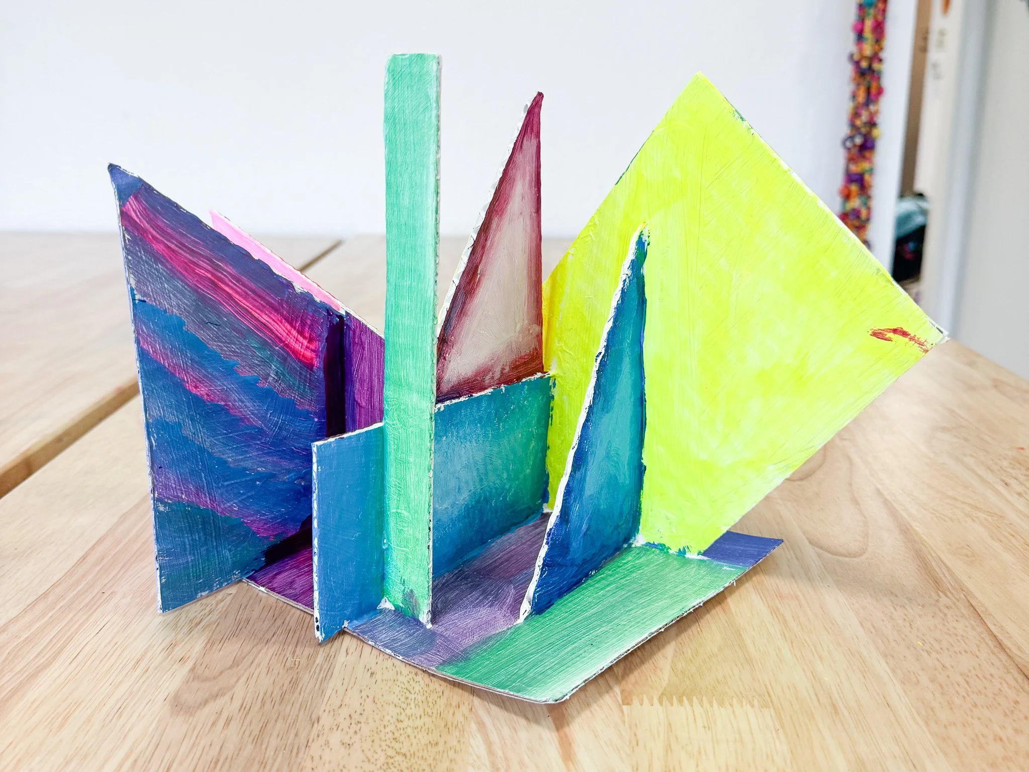 Colorful abstract painted cardboard structure with vertical and diagonal panels on a wooden table.