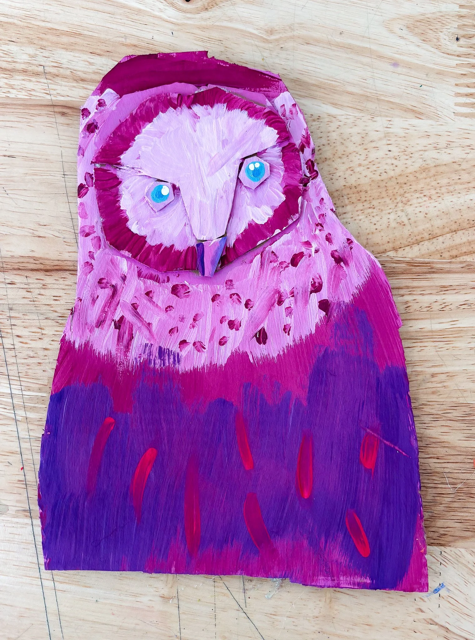 Painted picture of an owl with pink and purple colors, featuring blue eyes on a wooden surface.
