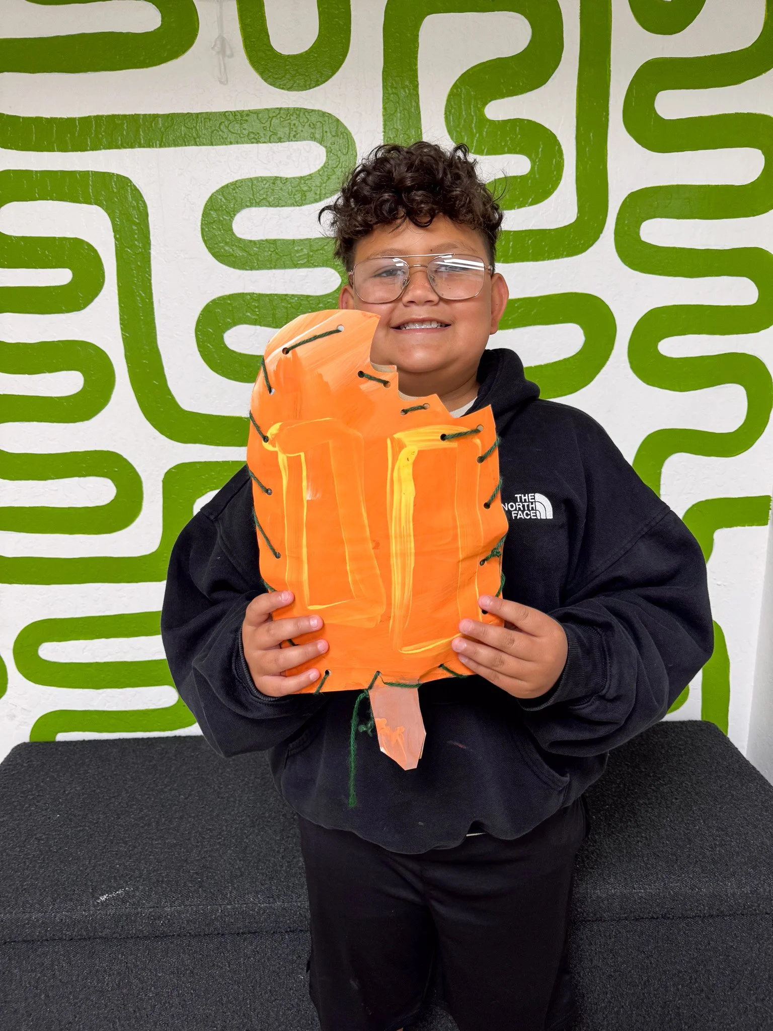 A smiling boy holding a painted paper pumpkin craft, standing in front of a green maze-like wall.