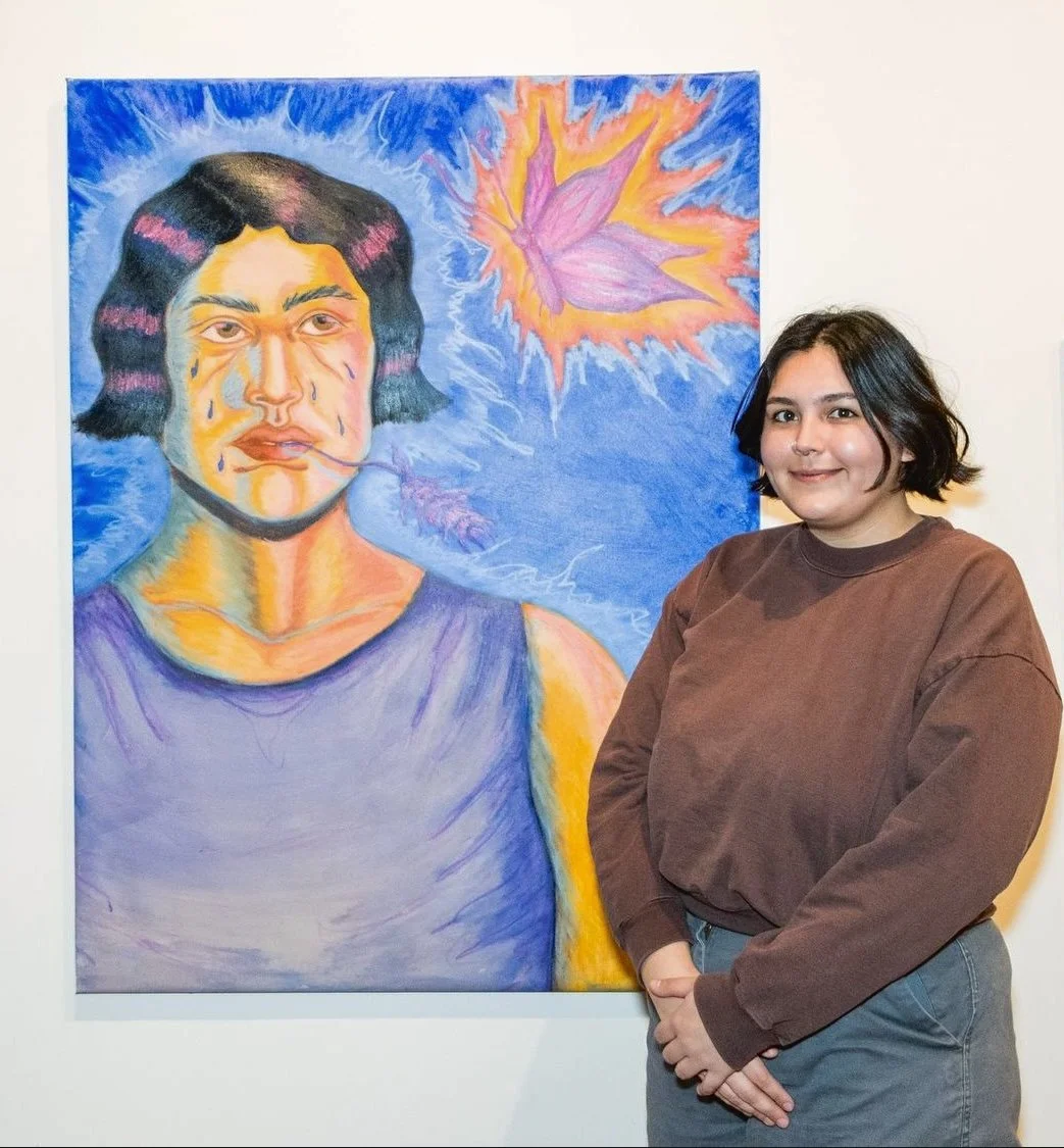 A young woman with short dark hair and a brown sweatshirt standing next to a colorful abstract portrait of a person with a blue background and a pink leaf with lightning around it.