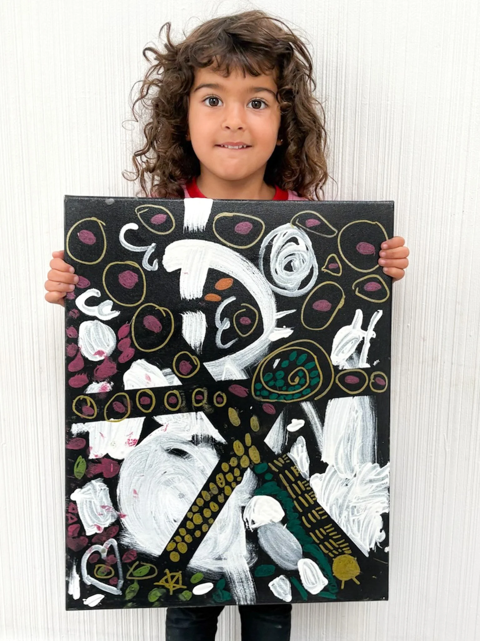 Young girl with curly hair holding a colorful abstract painting on canvas, featuring black background, white swirls, and various shapes in pink, green, and yellow.