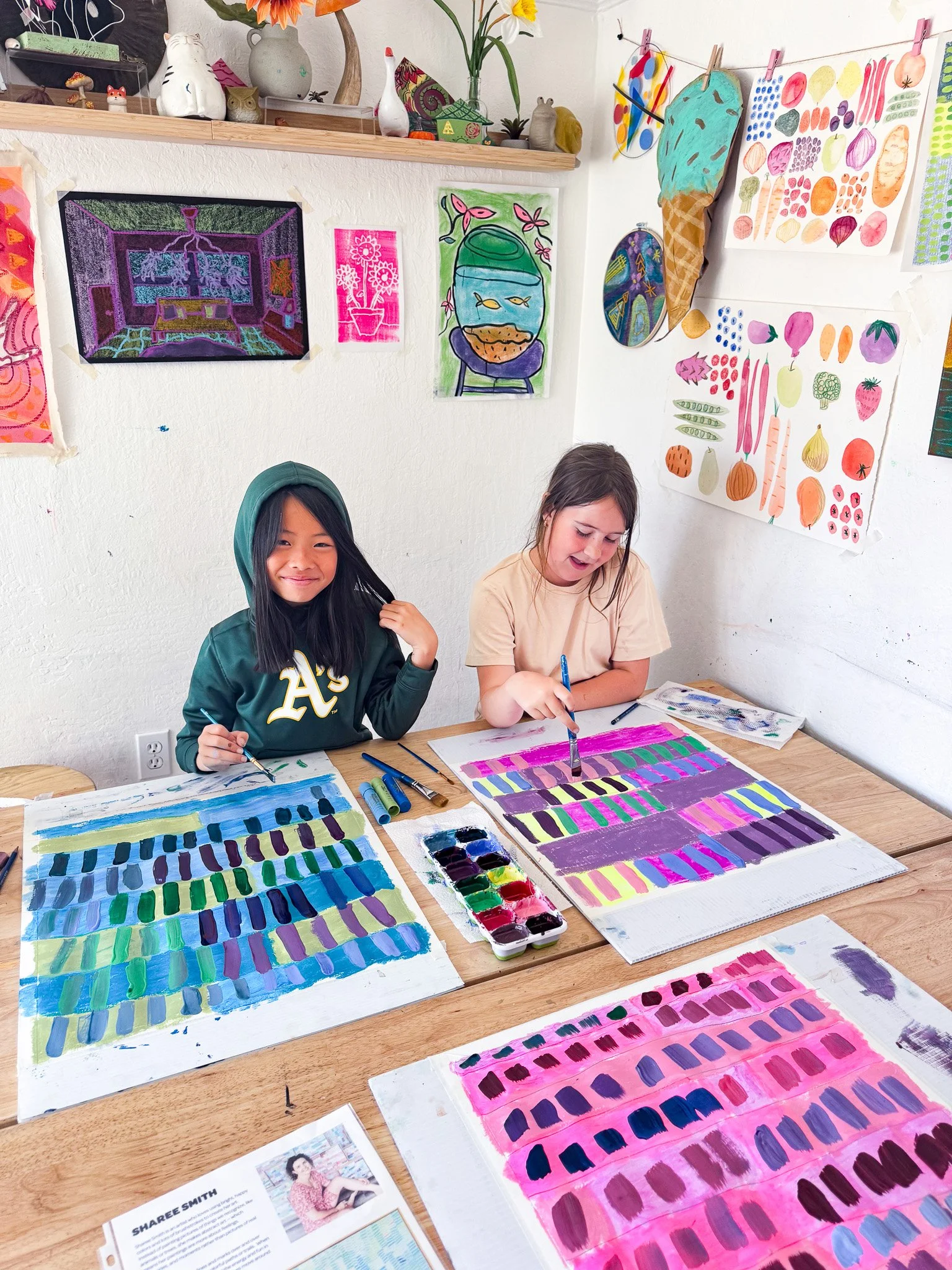 Two young girls are painting colorful abstract patterns on large sheets of paper at a wooden table in an art classroom. The room is decorated with various artworks and ceramics on the walls and shelves.