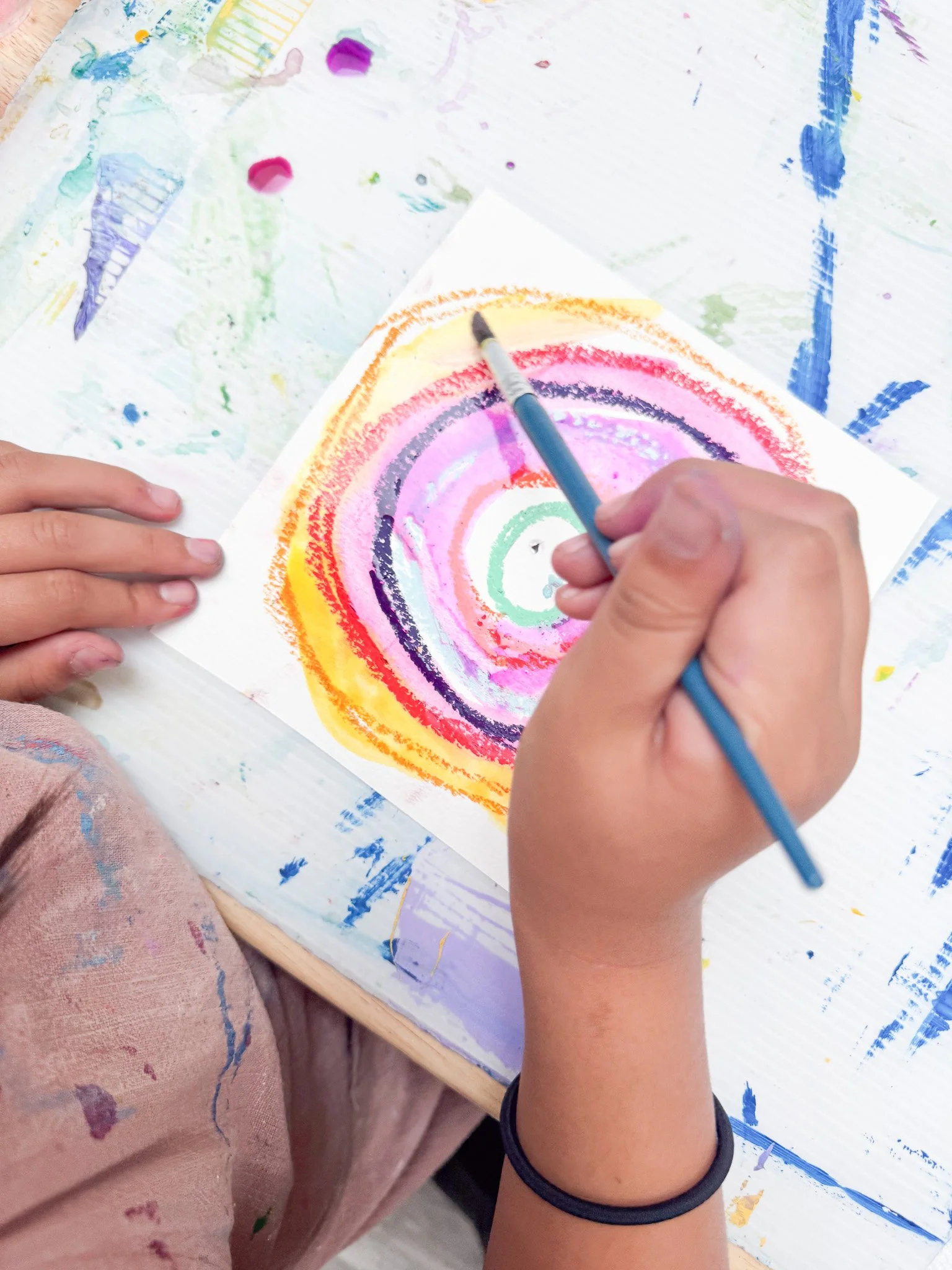 Child creating a colorful spiral painting with watercolors on a white sheet of paper, surrounded by a paint-covered table.
