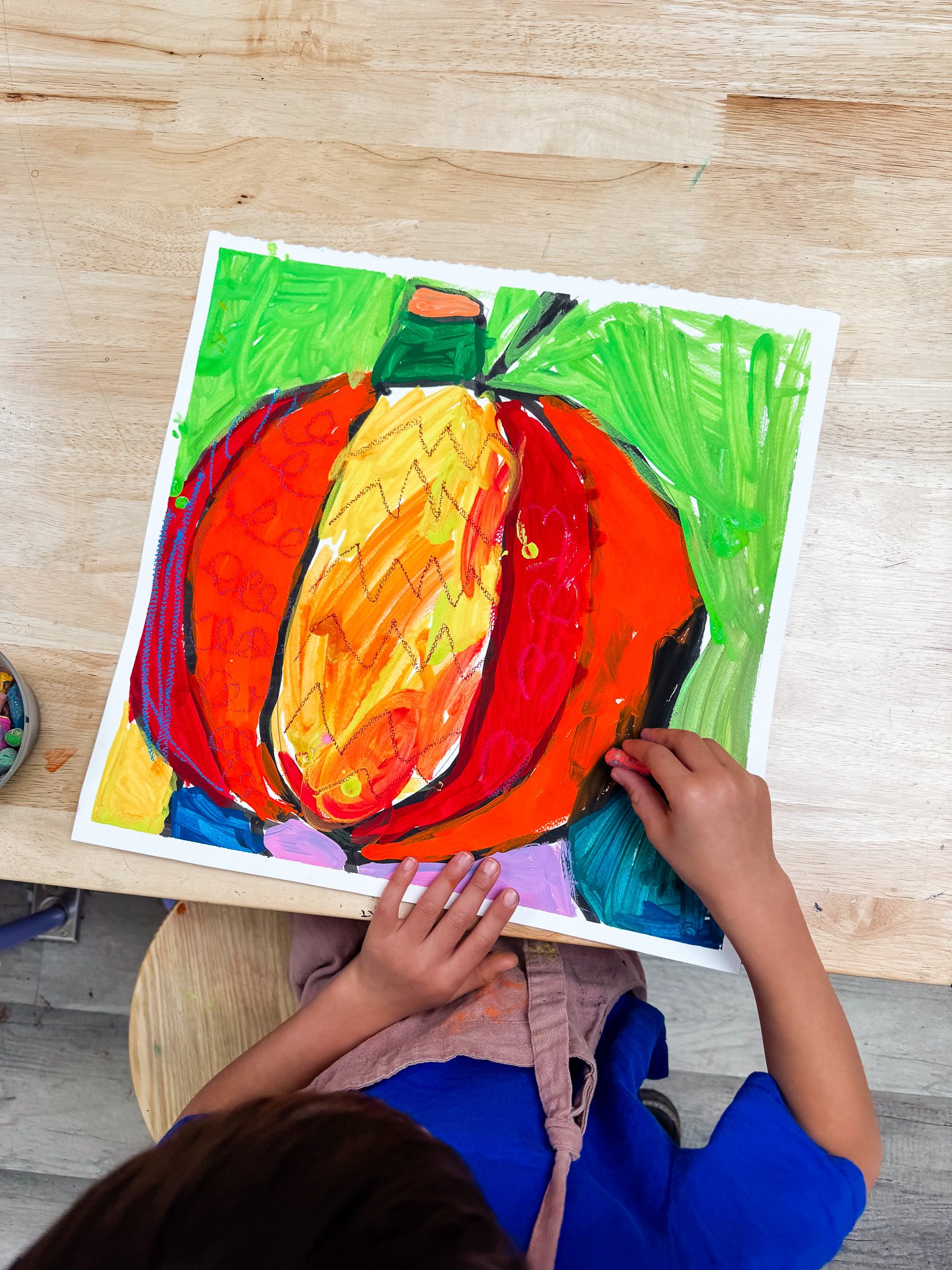 A child's colorful drawing of a pumpkin on paper, with green and purple background, placed on a wooden table. The child wears a blue shirt and beige shorts, with their hand touching the drawing.