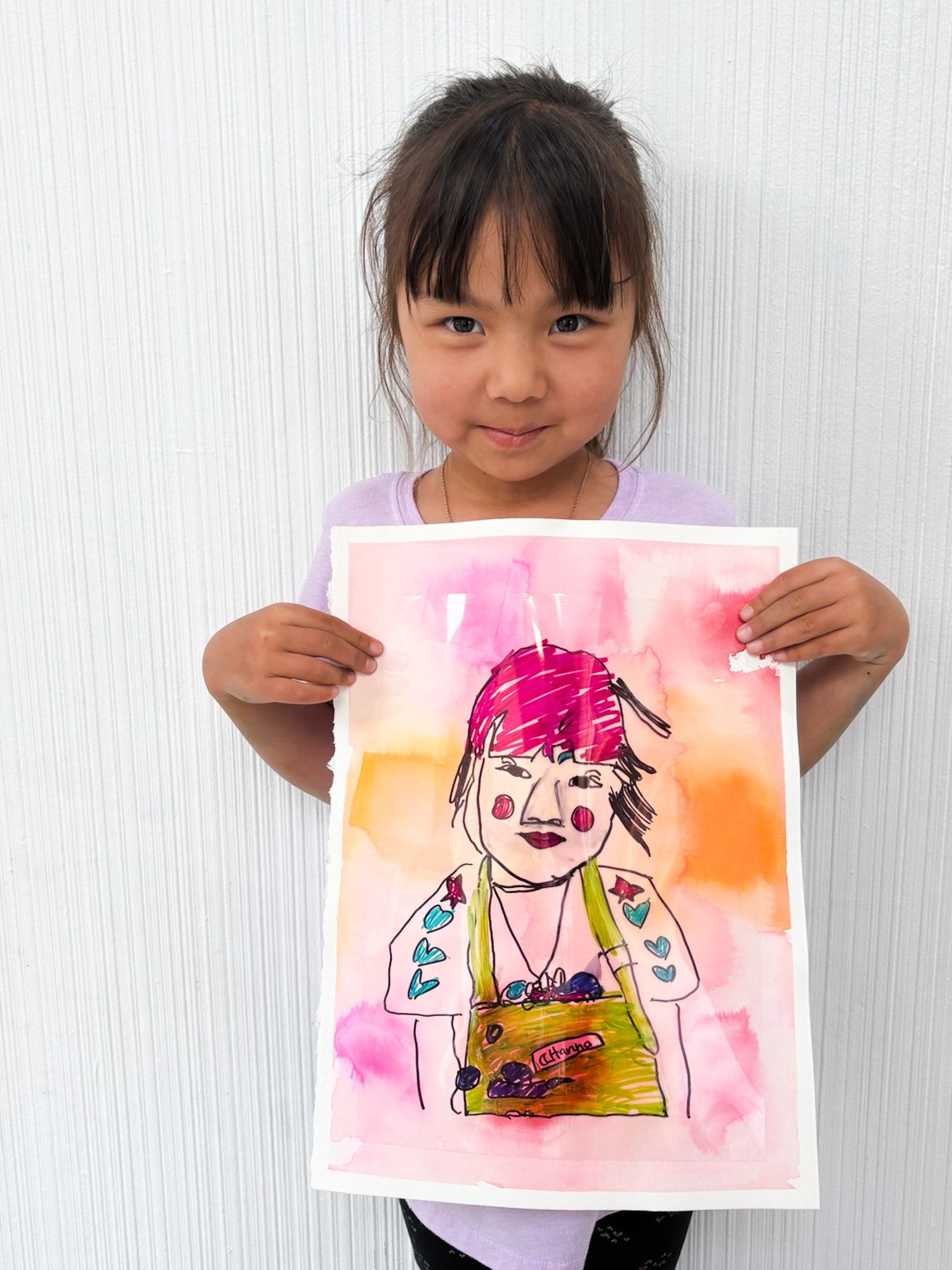 Young girl holding a colorful watercolor and ink drawing of a woman with pink hair, makeup, and a necklace, standing against a white wall background.