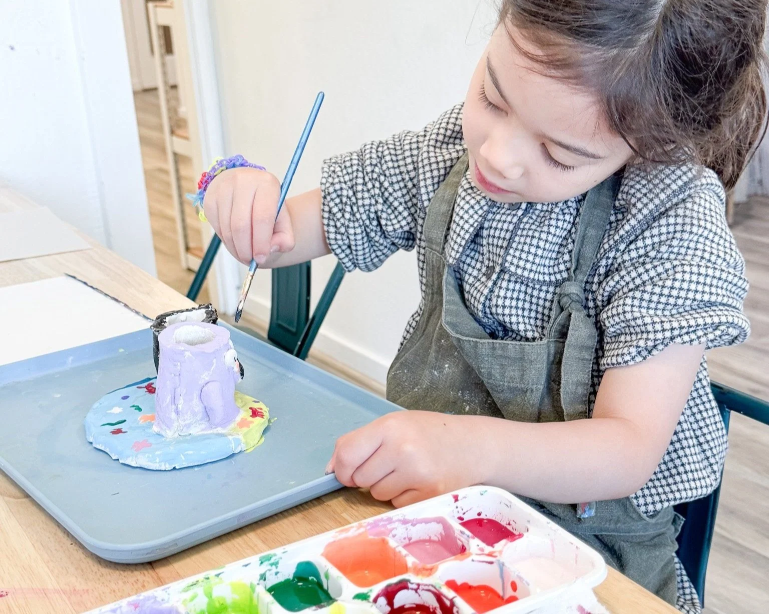 Young girl painting a ceramic sculpture with purple paint at a craft table.
