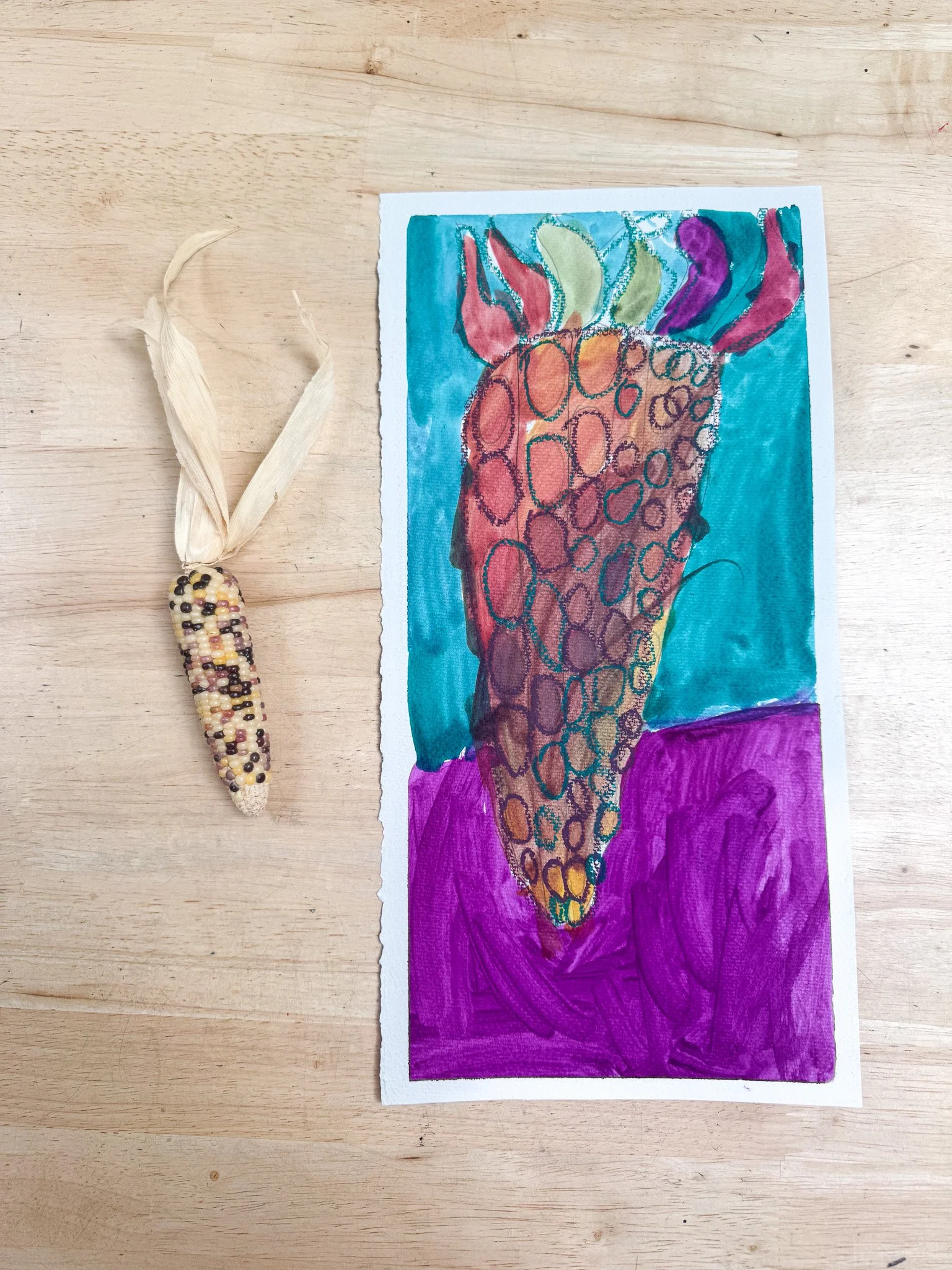 A colorful hand-painted illustration of a strawberry on paper with a small decorative object resembling a tiny corn on the cob made of beads to the left, both placed on a wooden surface.