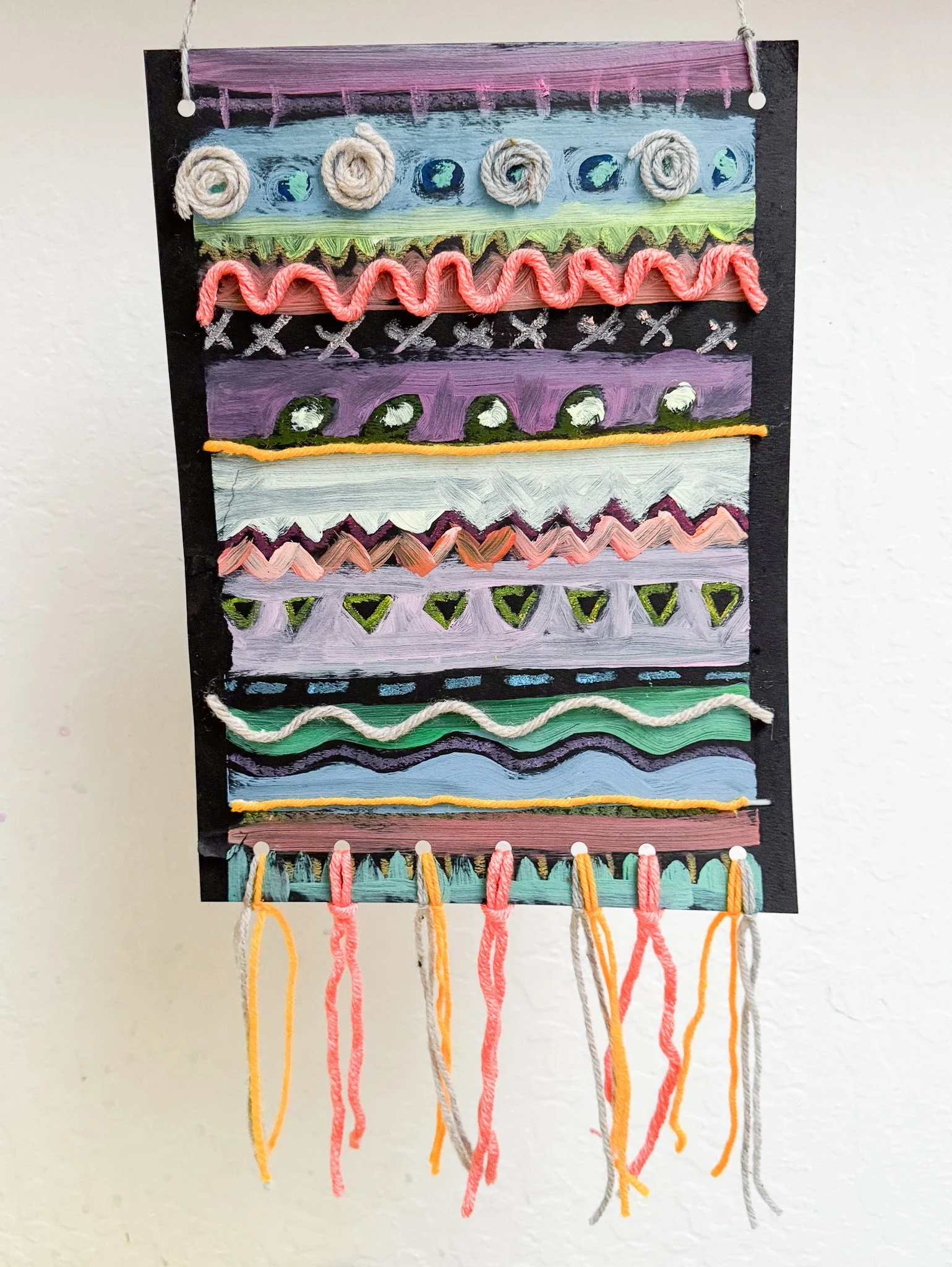 Colorful yarn and paint patterns on a black hanging board with strings of yarn hanging at the bottom.