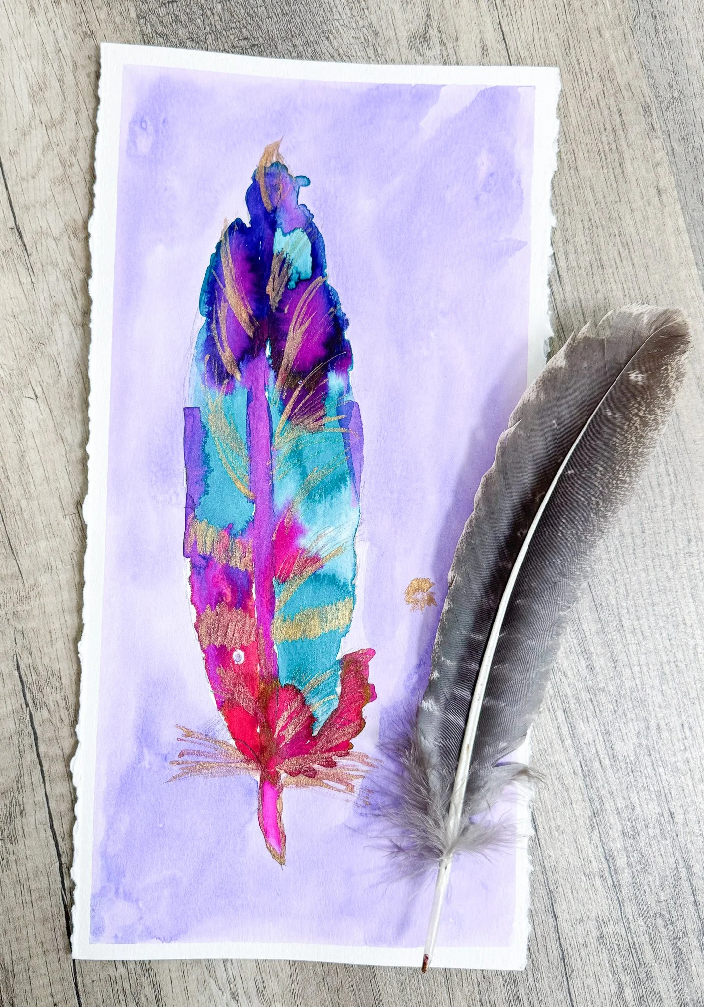 A colorful watercolor illustration of a feather placed on a light purple background, with a real brown and white feather resting beside it on a wooden surface.