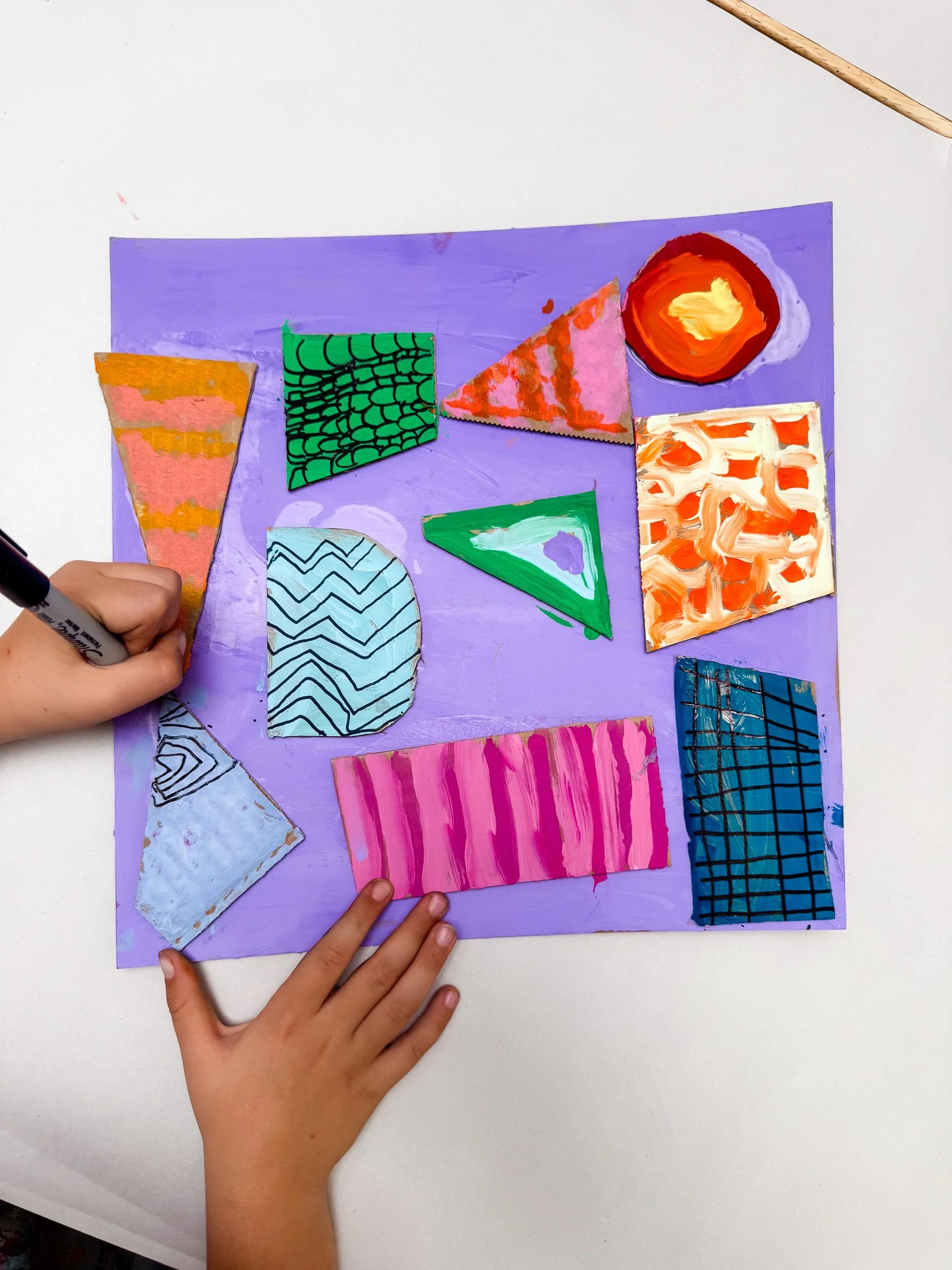 Children's hands working on abstract painted paper pieces arranged on a purple background.