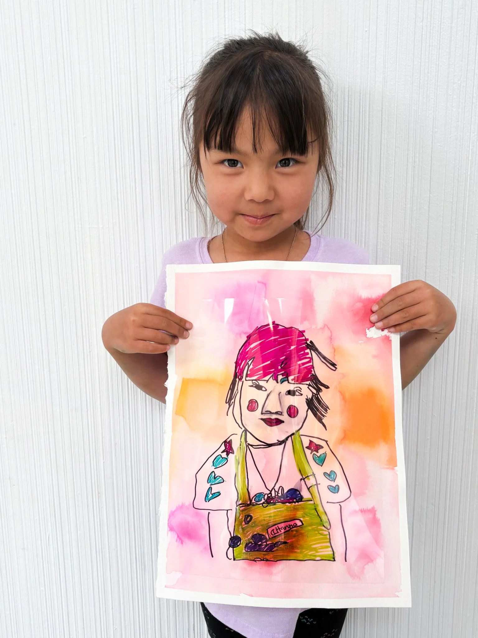 Young girl holding a colorful watercolor and ink portrait of herself, standing against a white textured background.