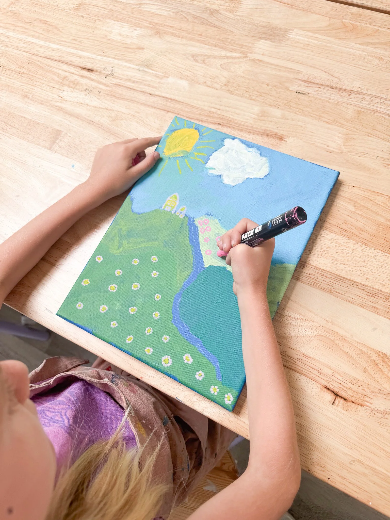 Child painting a landscape on a canvas, depicting the sun, a cloud, rolling green hills, a blue river, and flowers.