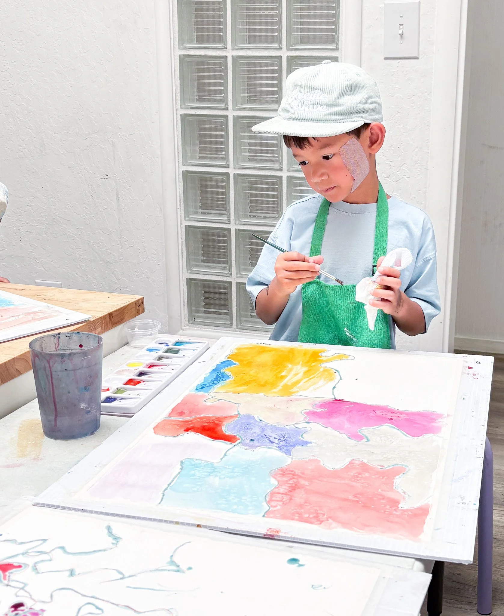 Young boy painting on a canvas with vibrant colors, wearing a light blue shirt, a white hat, and a green apron, in an art studio.
