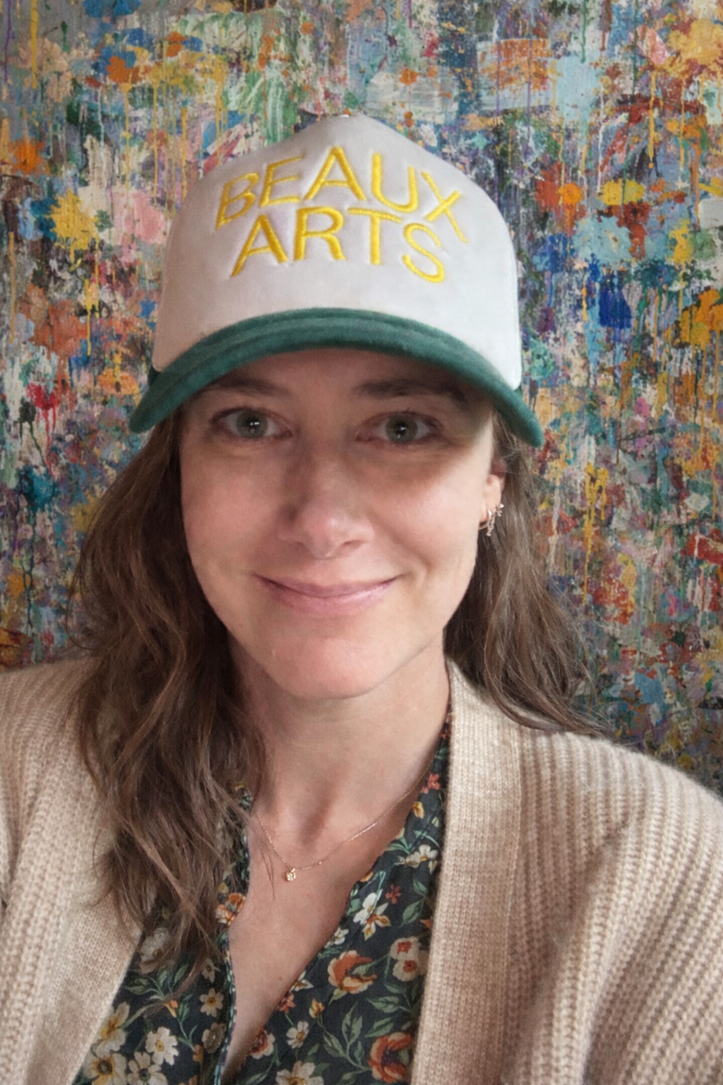 A woman wearing a beige cardigan and floral shirt, smiling in front of an abstract, colorful splattered paint background, with a white cap that says 'BEAUX ARTS' in yellow lettering.