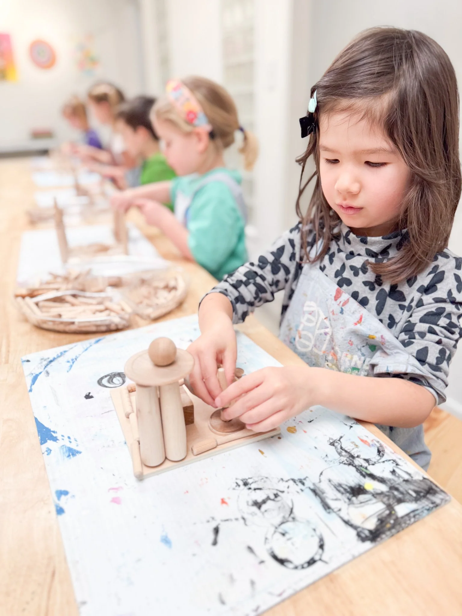 A young girl in a patterned shirt working with wooden toys at a table in a classroom with other children in the background.