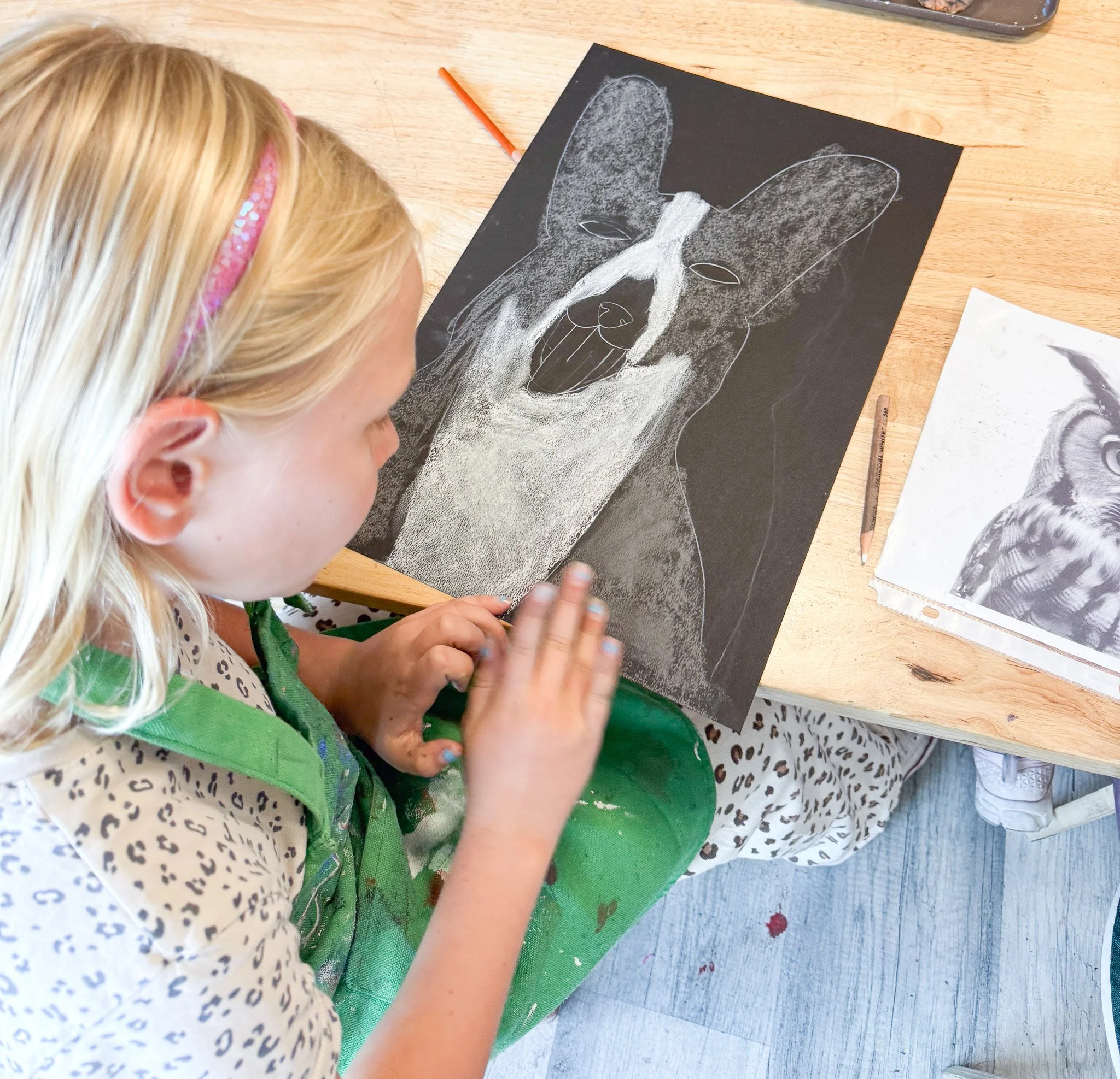 A young girl in a green apron and leopard print pants is creating a drawing of a dog's face on black paper using white chalk. She is focused on her artwork, with a reference photo of a dog on the table beside her.