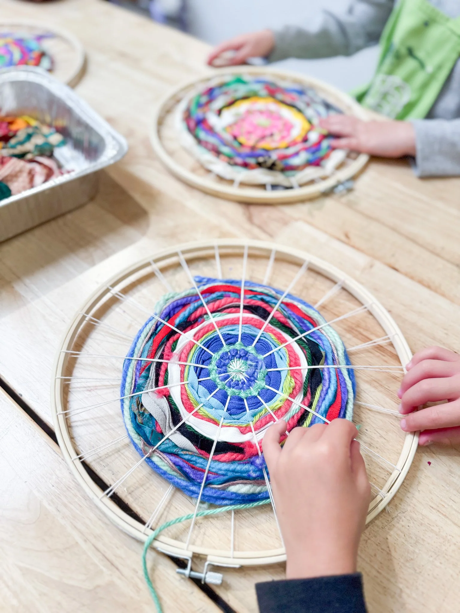 Child creating a woven circular art piece using colorful fabric and a small embroidery hoop on a wooden table.