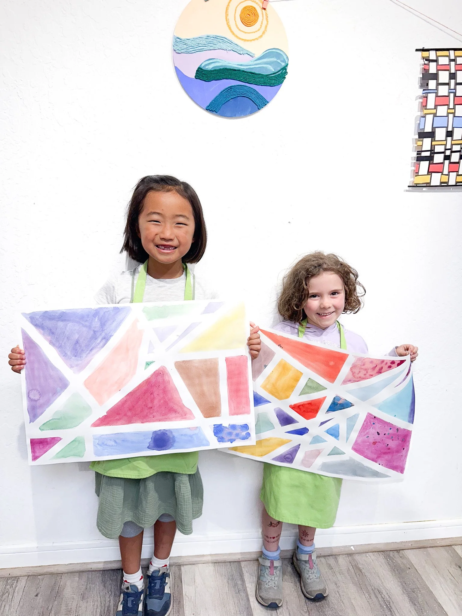 Two young girls are standing in front of a white wall, each holding a colorful abstract painting. The girl on the left has dark hair and is smiling, wearing a white shirt and a green skirt. The girl on the right has curly hair and is also smiling, we