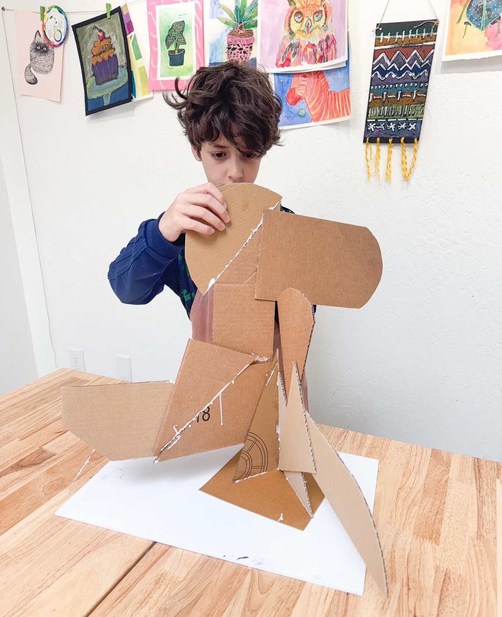 A young boy with curly hair wearing a dark long-sleeve shirt building a sculpture from large cardboard pieces at a wooden table. The cardboard sculpture resembles an abstract humanoid figure.