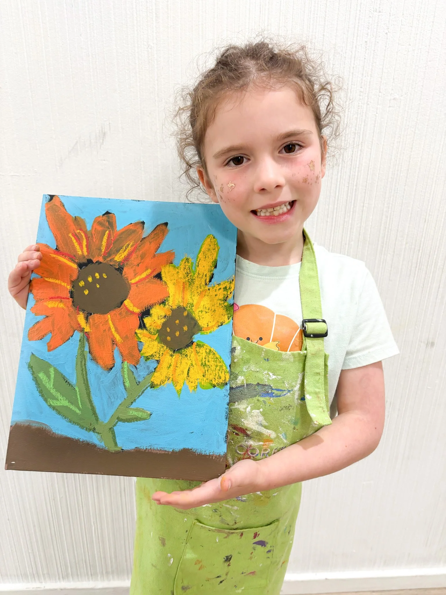 A young girl with curly hair holding a painting of two colorful flowers, one orange and one yellow, against a light blue background.