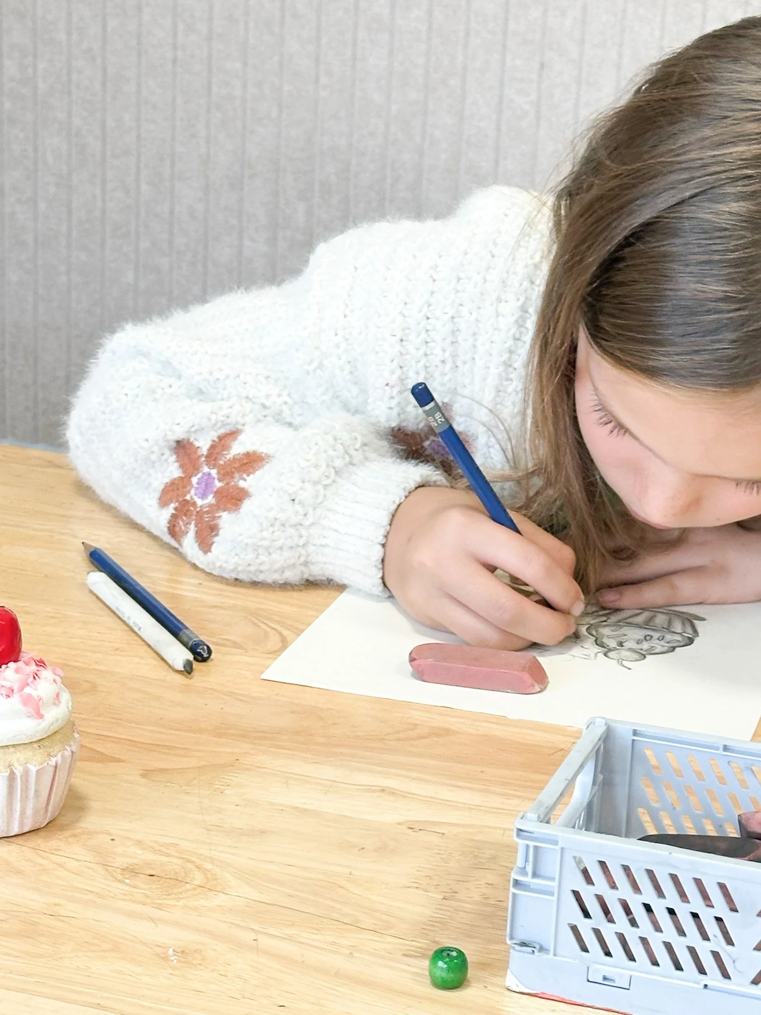 Young girl drawing with colored pencils on paper at a wooden table, with a cupcake, a pen, and a green marble nearby.