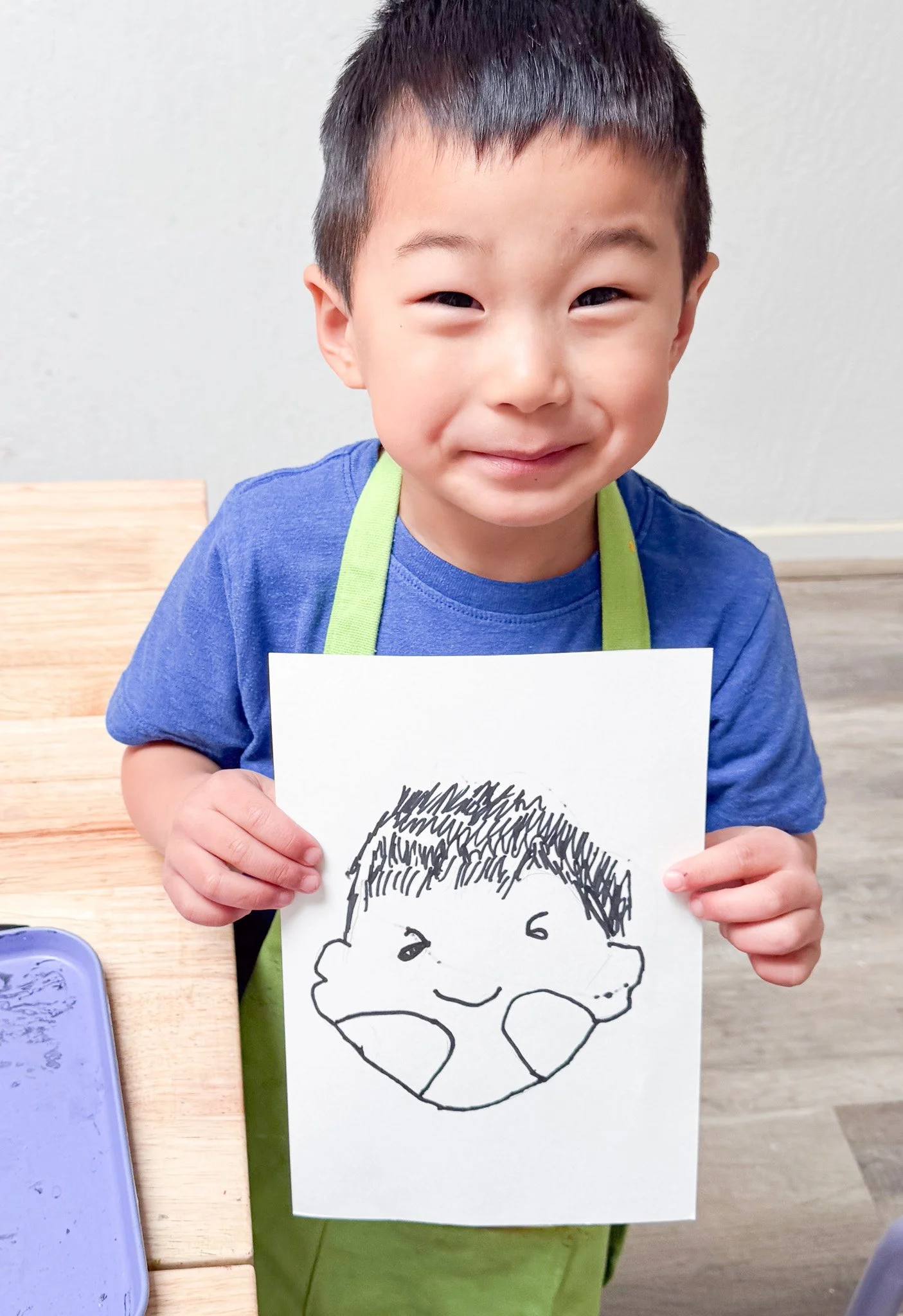 A young boy holding a drawing of a smiling face with a head of hair and big cheeks, smiling at the camera.