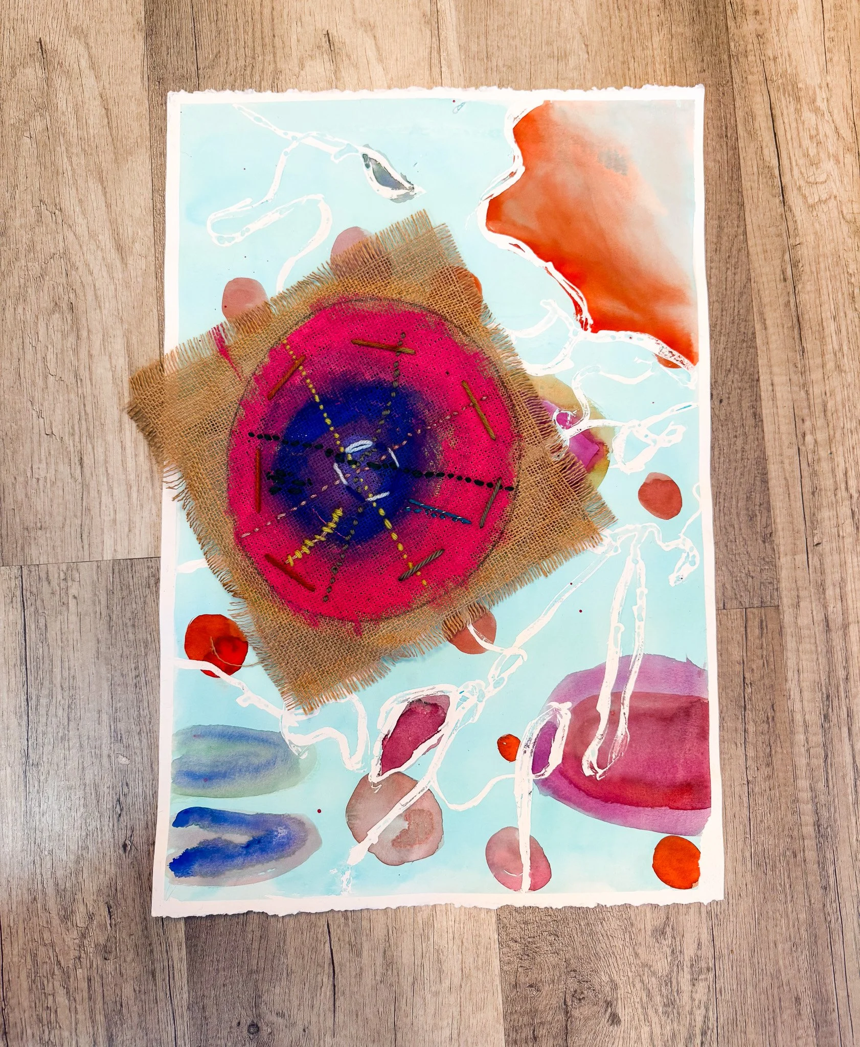 Abstract mixed-media artwork featuring a central circular design with vibrant pink, purple, and blue colors, on fabric with frayed edges, surrounded by watercolor shapes and splatters in red, orange, blue, and beige on paper.