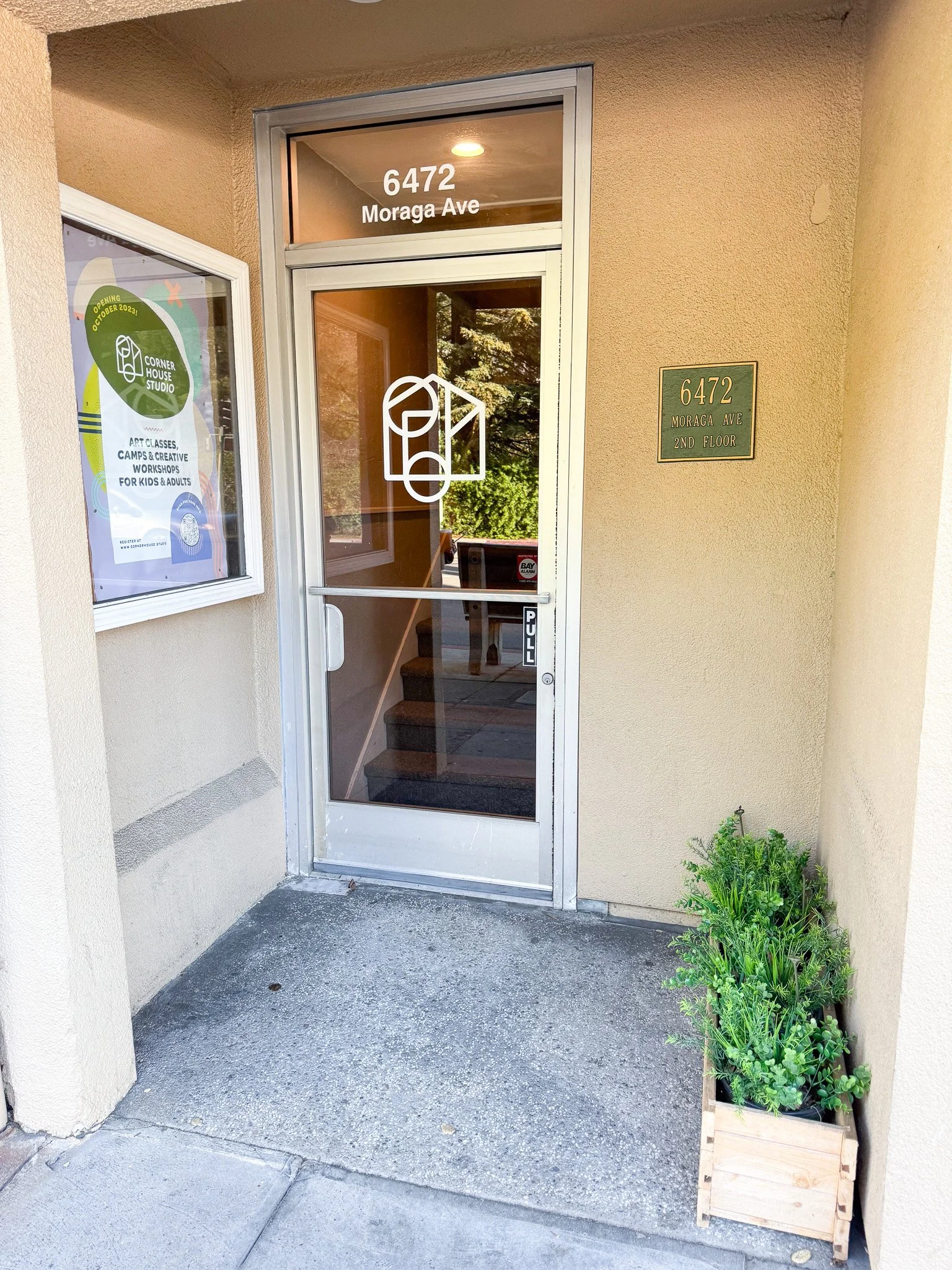 The entrance to a building with a glass door displaying the address '6472 Moraga Ave' and a sign indicating the second floor. To the left, there is a window with a poster advertising art classes and workshops for kids and adults at Corner House Studi