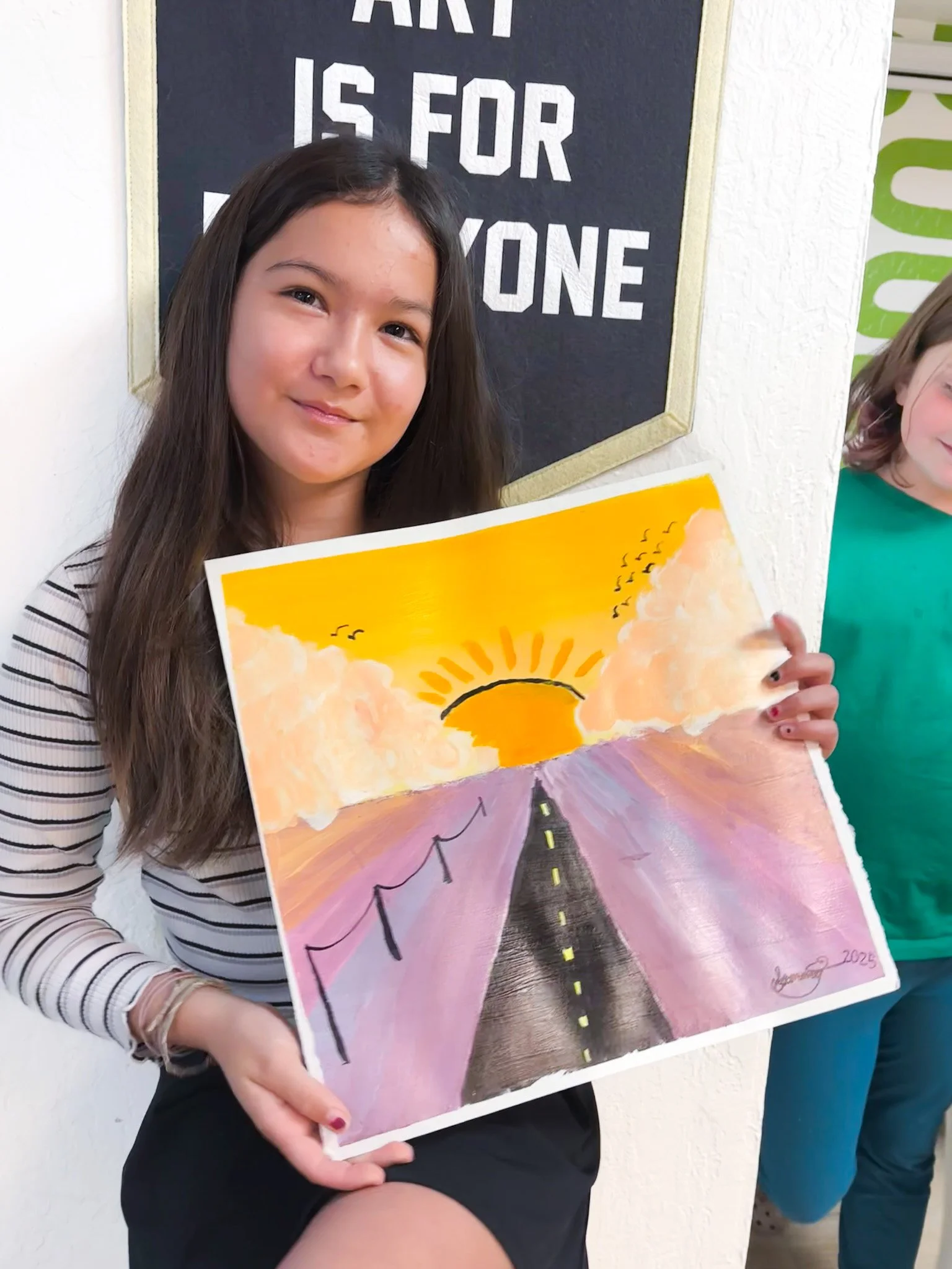 A young girl holding a colorful painting of a road leading to a sunset with clouds and birds, standing in front of a black poster with white text.