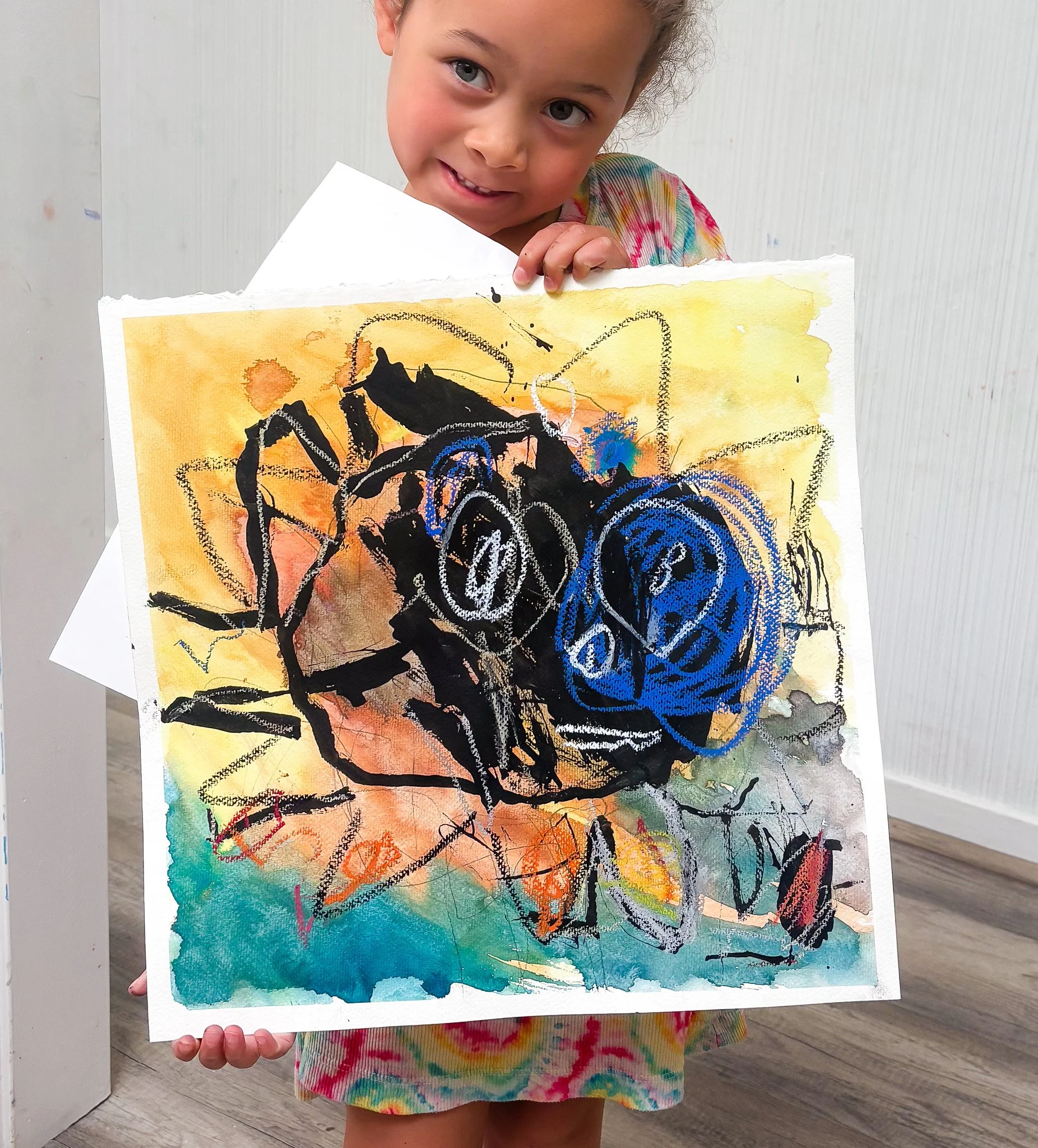 A young girl with brown hair in a ponytail holding up a colorful abstract painting she created, smiling at the camera, in an indoor setting.