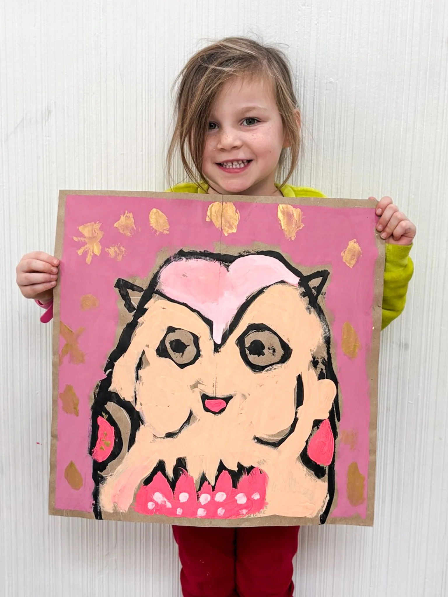 Young girl smiling and holding a colorful painting of a cartoon-style dog with pink and beige fur, surrounded by gold and pink accents on a pink background.