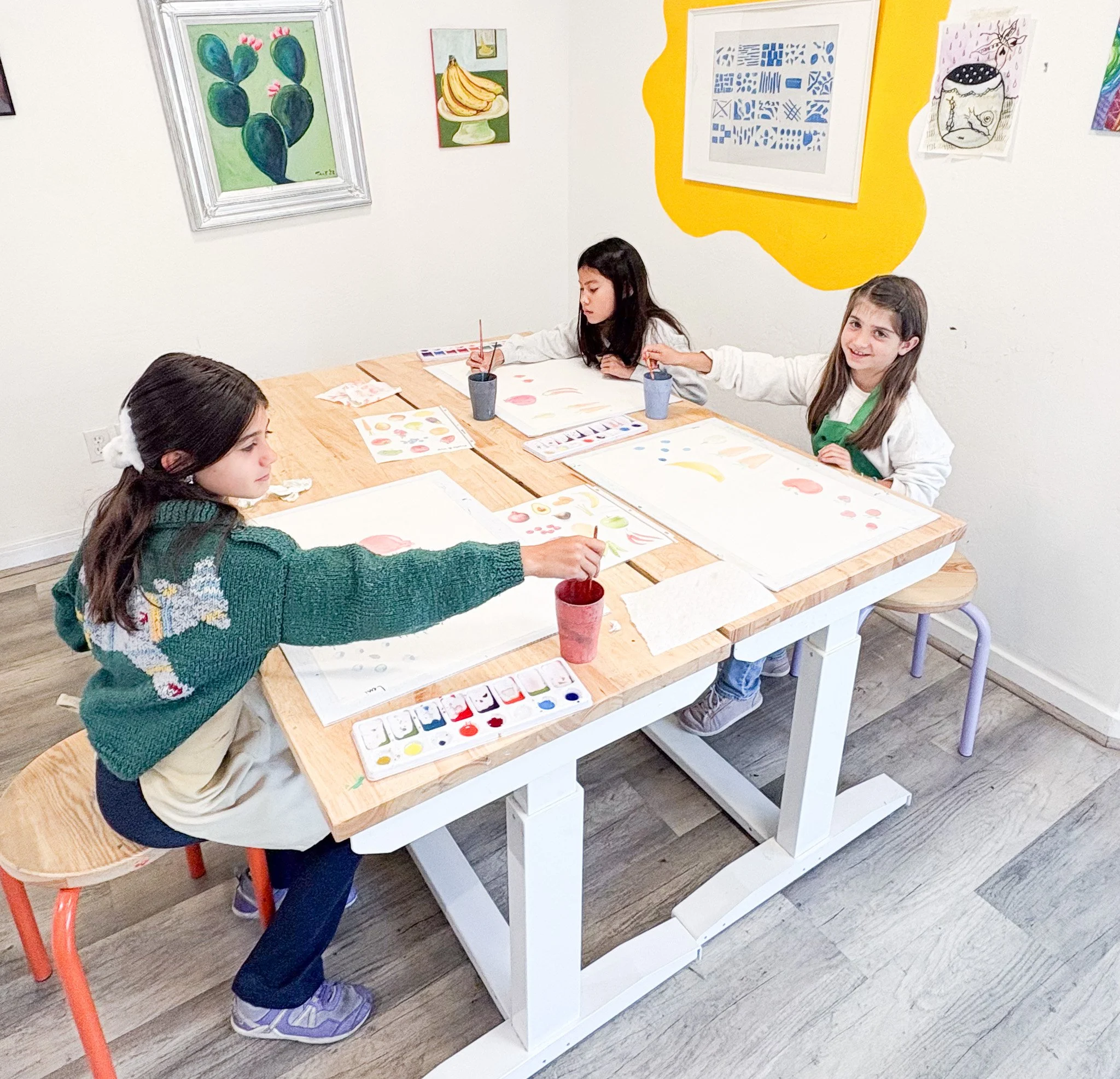 Three young girls are painting watercolor pictures at a wooden table in an art classroom. They are surrounded by colorful artwork on the walls.