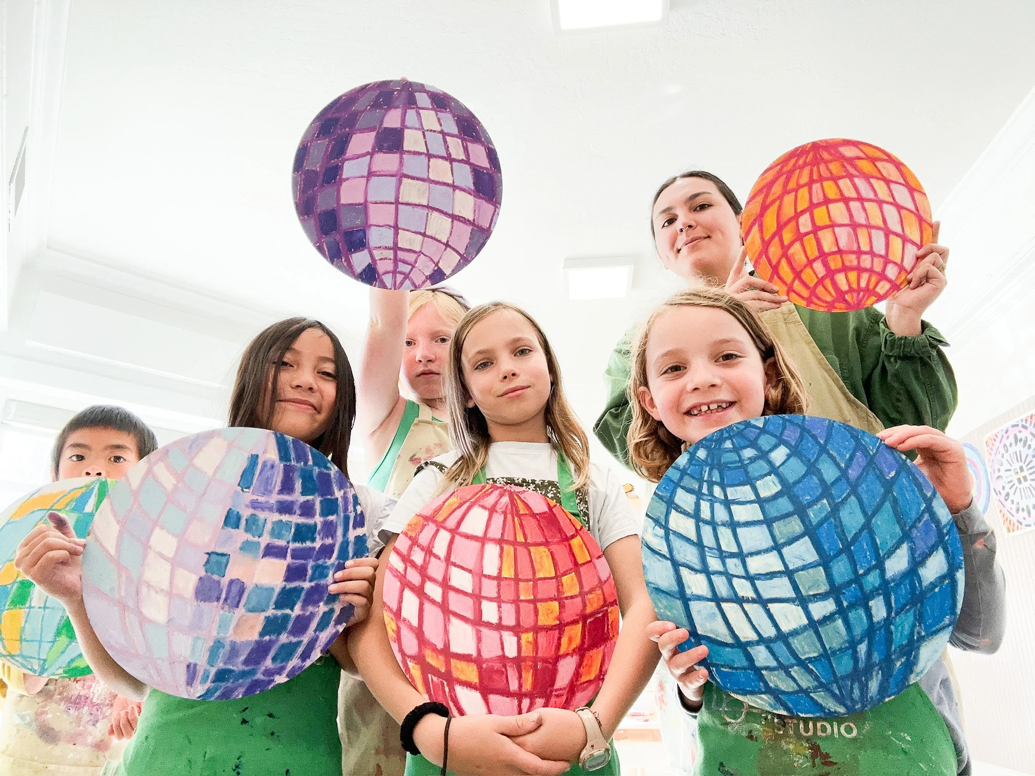 Children and a woman holding colorful, painted paper lanterns with geometric patterns in a bright room.