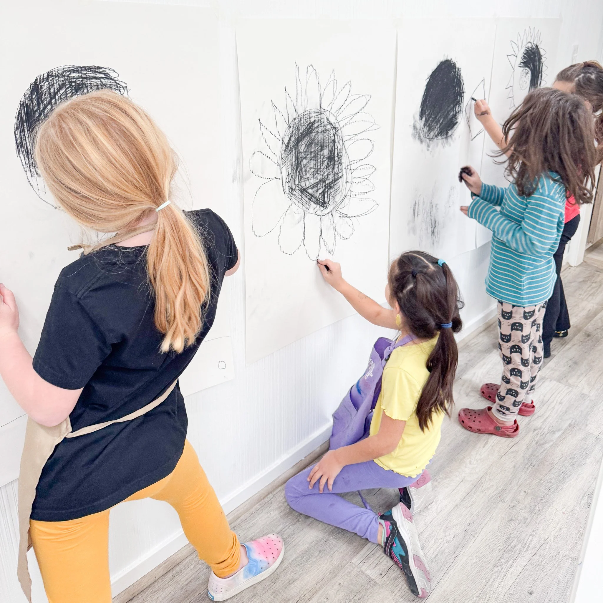 Children drawing large sunflowers with black crayon on white paper displayed on the wall at an art class.