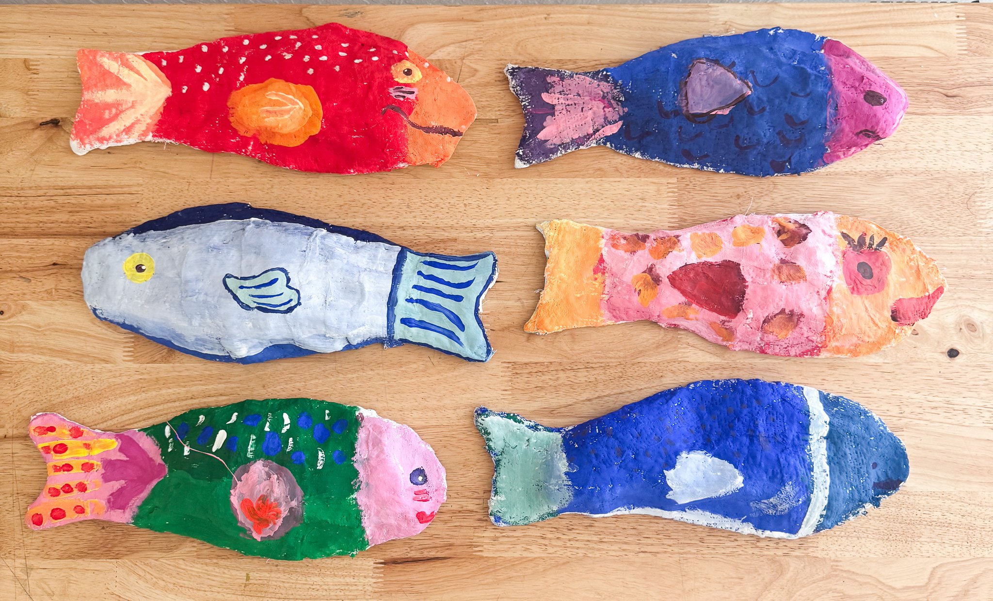 Six colorful, hand-painted paper fish crafts arranged on a wooden surface, each with unique colors and patterns.