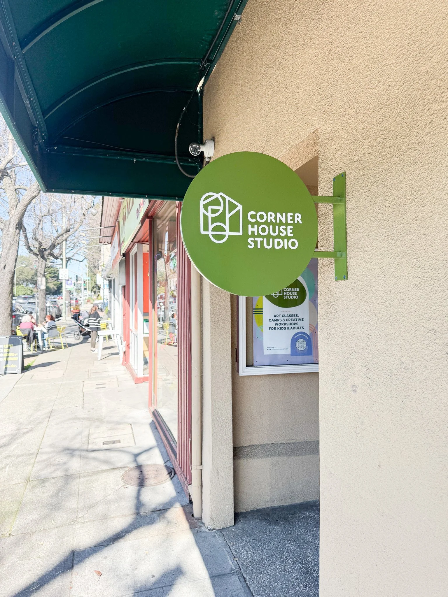 Green round sign with white text and logo reading 'Corner House Studio' mounted on a beige wall outside a storefront, with a sidewalk and outdoor seating visible.