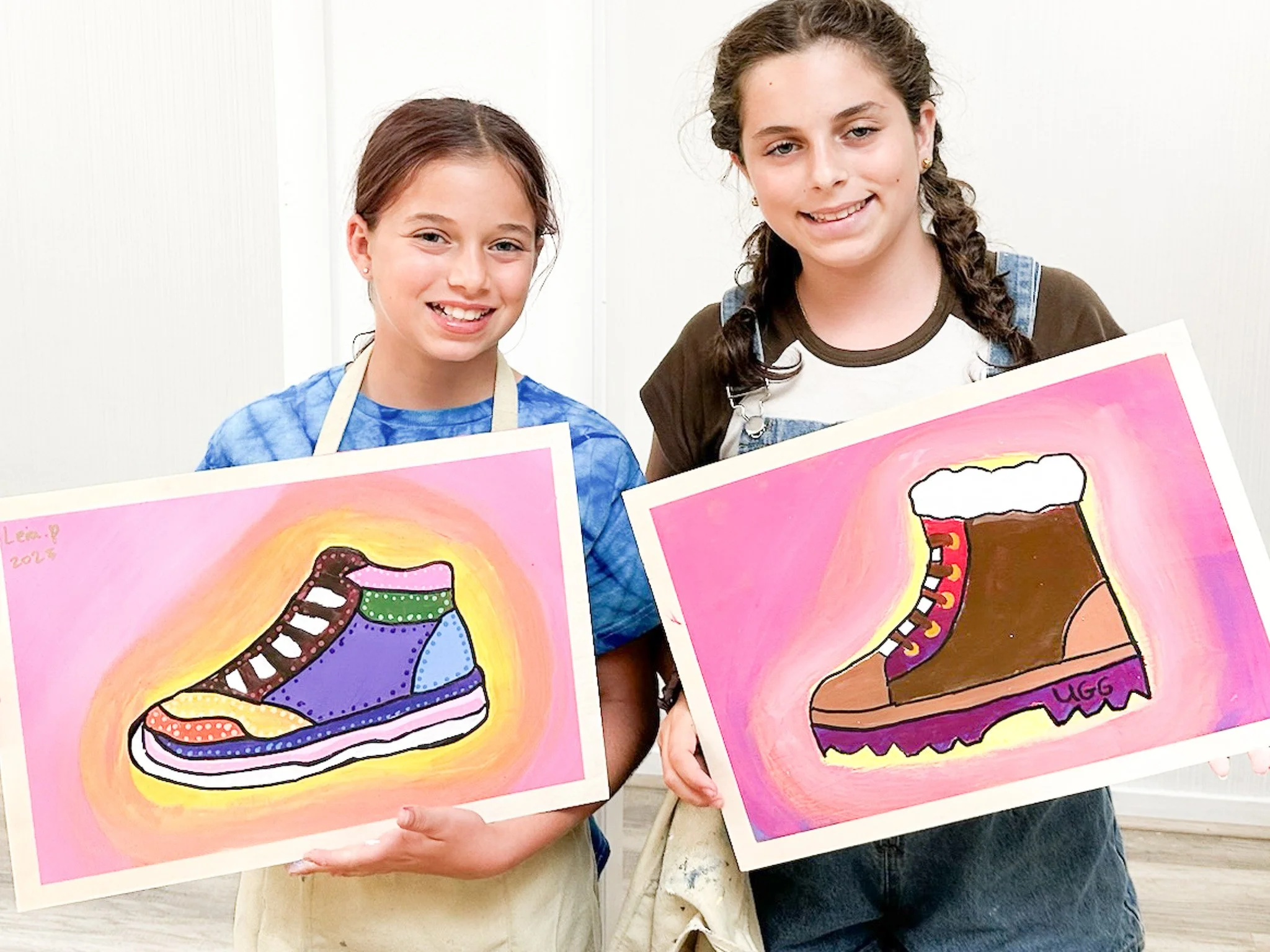 Two young girls holding colorful, cartoon-style paintings of children's shoes, standing indoors in front of a white wall.