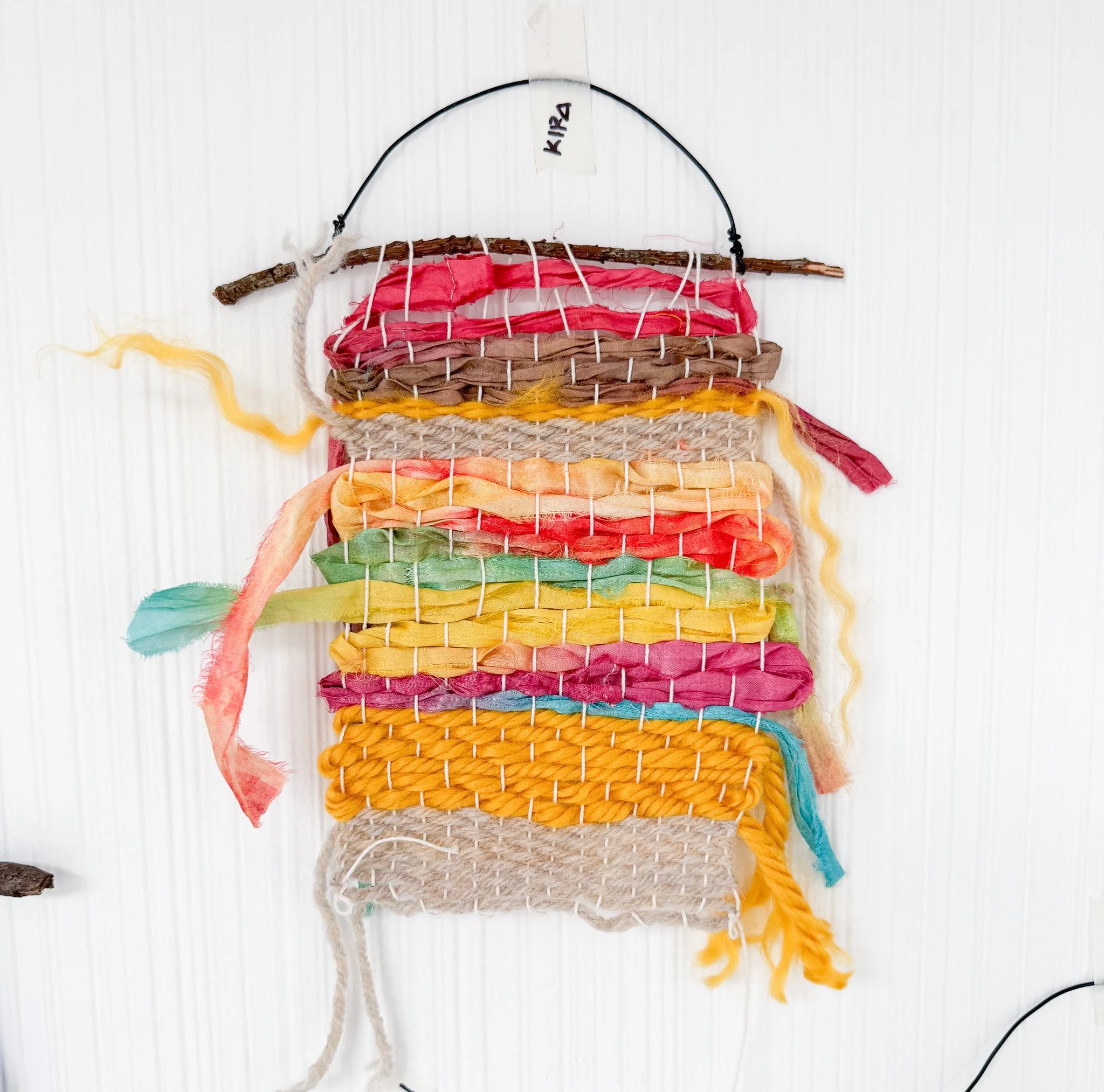 Colorful woven wall hanging made from fabric strips and sticks, with the name 'Kira' on a hanging loop at the top.