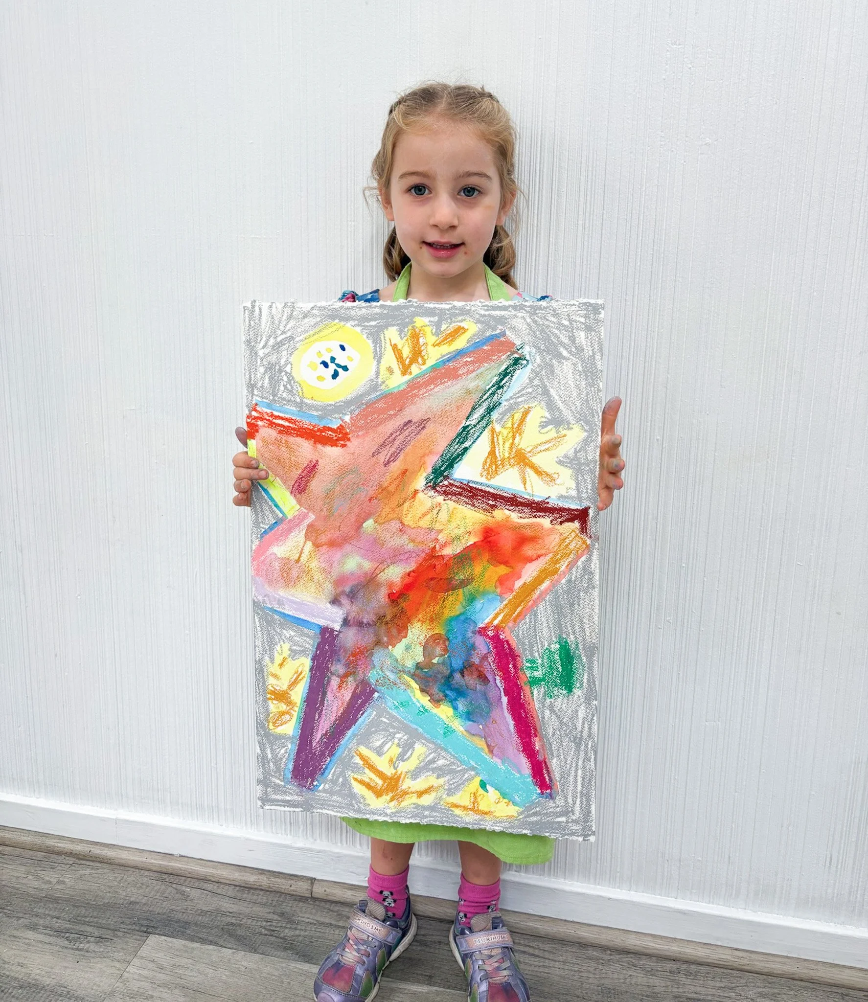 A young girl holding a colorful drawing of a star, with a rainbow, yellow sun, and other abstract shapes on white paper