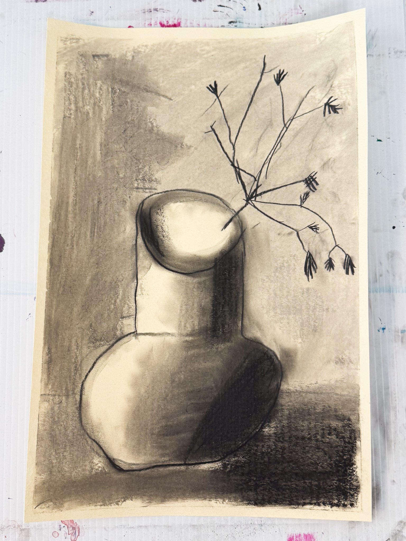 A charcoal drawing of a simple round vase with a rounded body and neck, holding a branch with small, sparse leaves and tiny flowers, placed on a flat surface with shadows cast to the right.