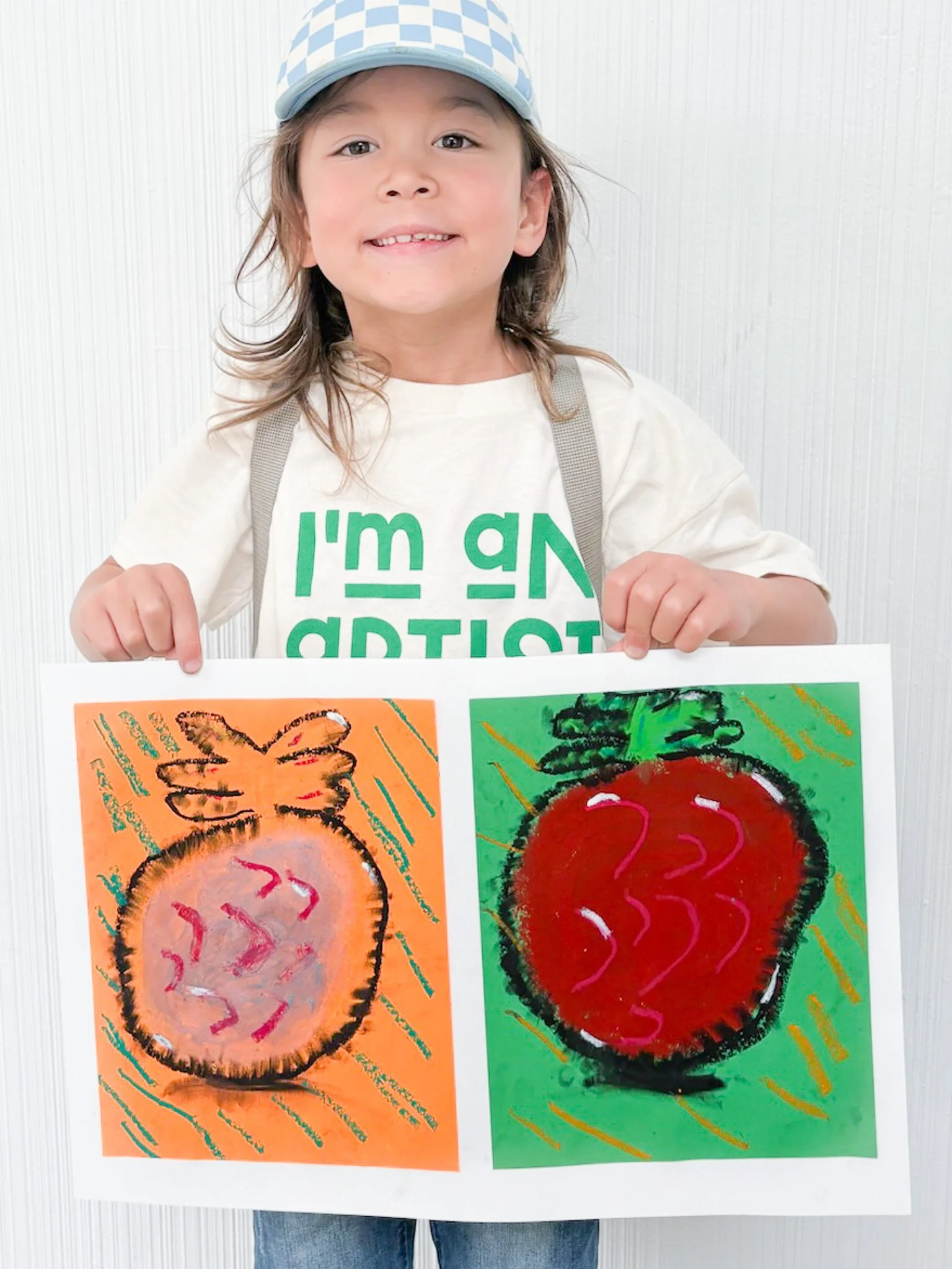 Young girl wearing a cap and an 'I'm an Artist' shirt, smiling and holding a colorful piece of artwork featuring two red strawberries on orange and green backgrounds.