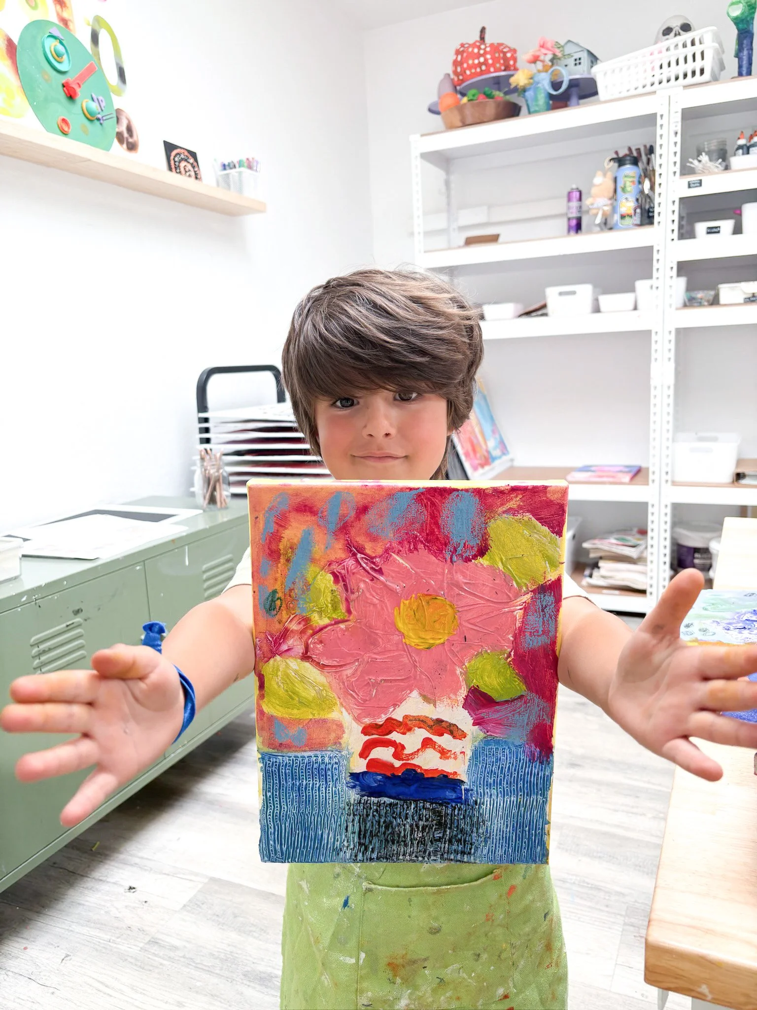 A young boy holding up a colorful, abstract painting in an art classroom.