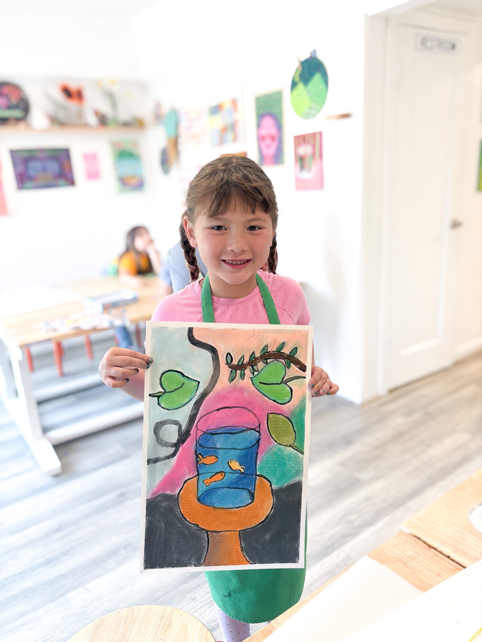 Young girl with braided hair wearing a pink shirt and green apron holding a colorful painting of a fish bowl on a stand, with a tree and leaves in the background, inside an art classroom.