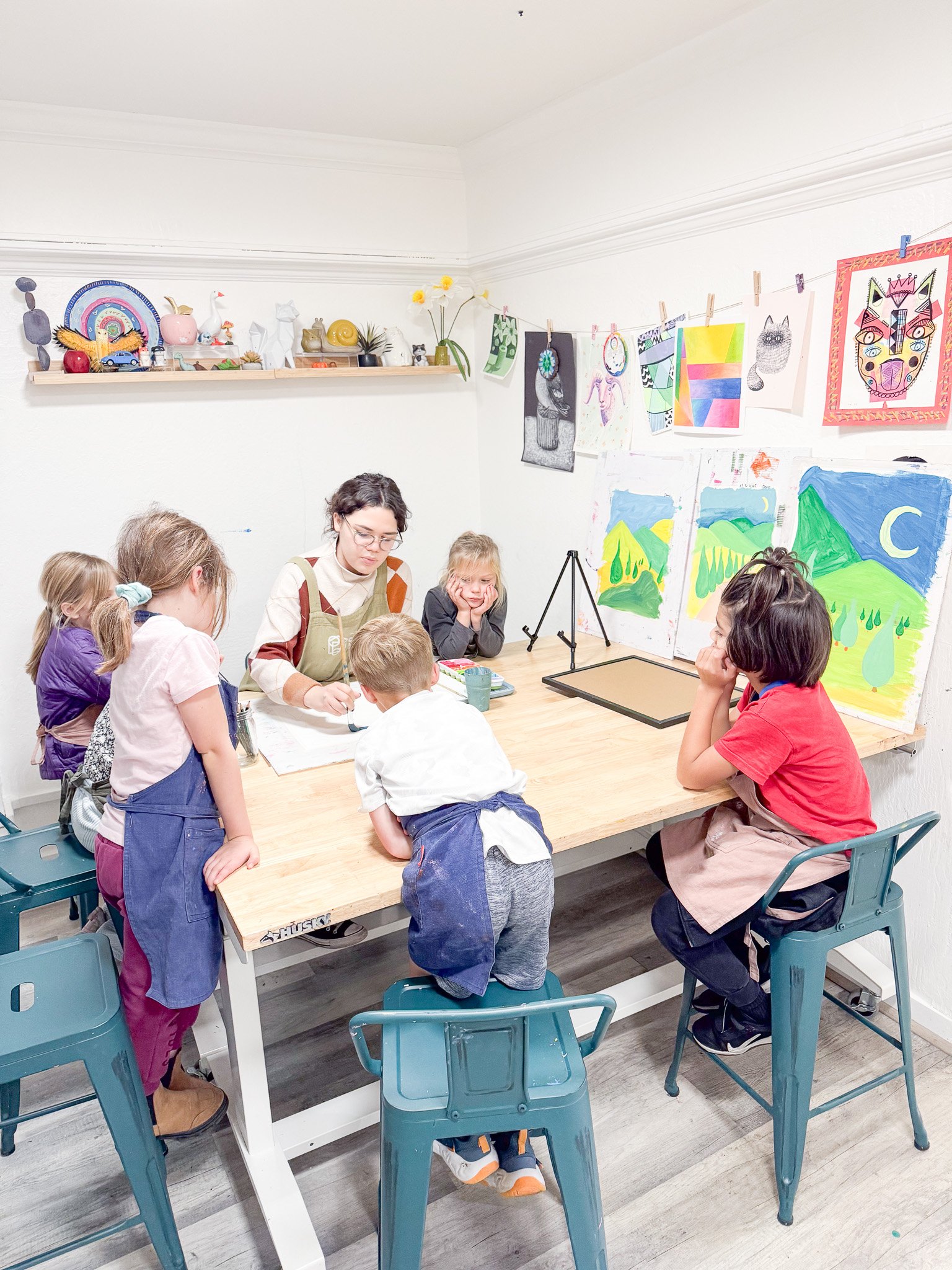 Children and an instructor sitting around a crafting table in an art classroom with colorful artwork and decorations on the walls.